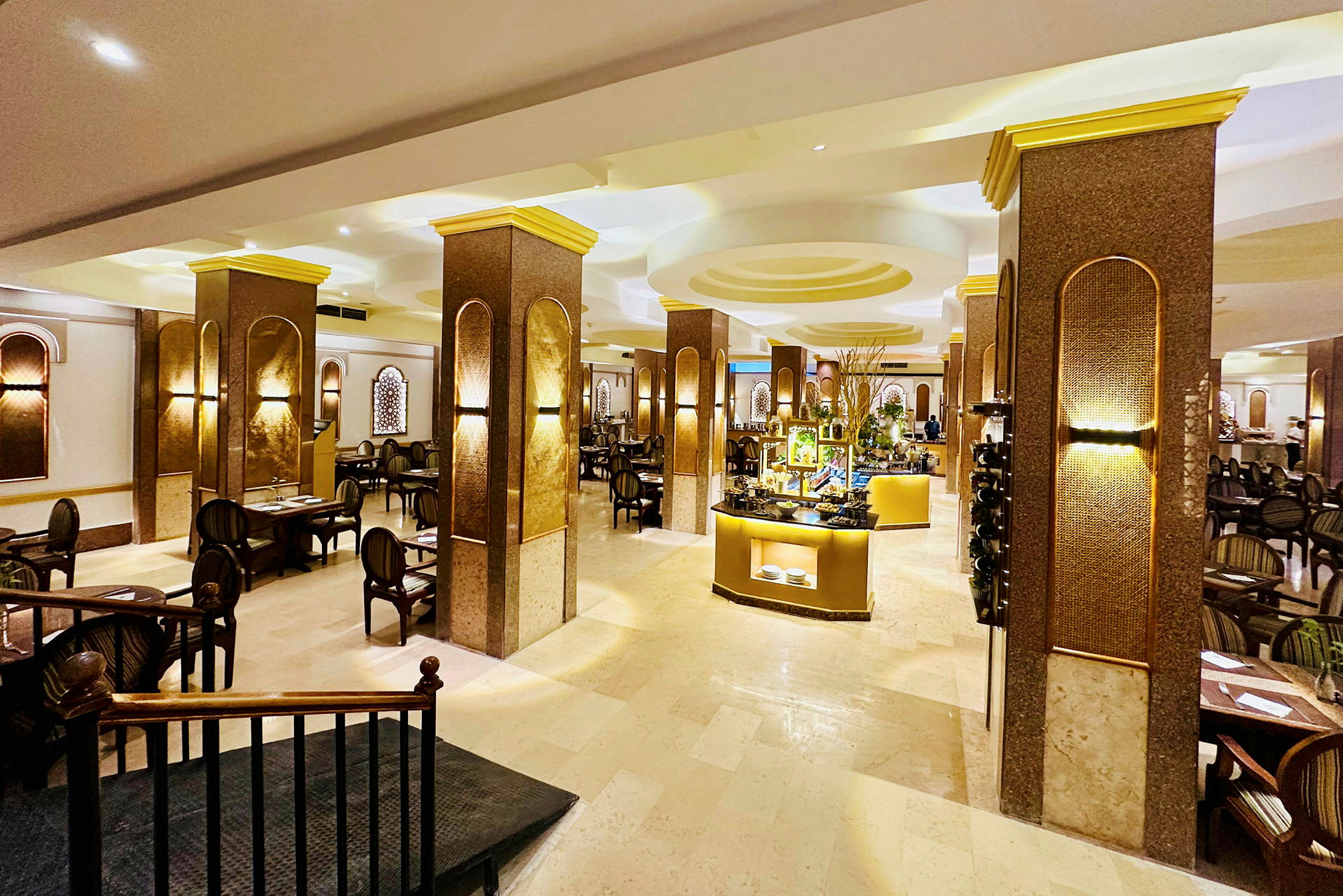 Et interiør af en elegant restaurant med borde og stole placeret mellem søjler. I midten er en buffet med belyste madstationer. (AI-genereret tekst)