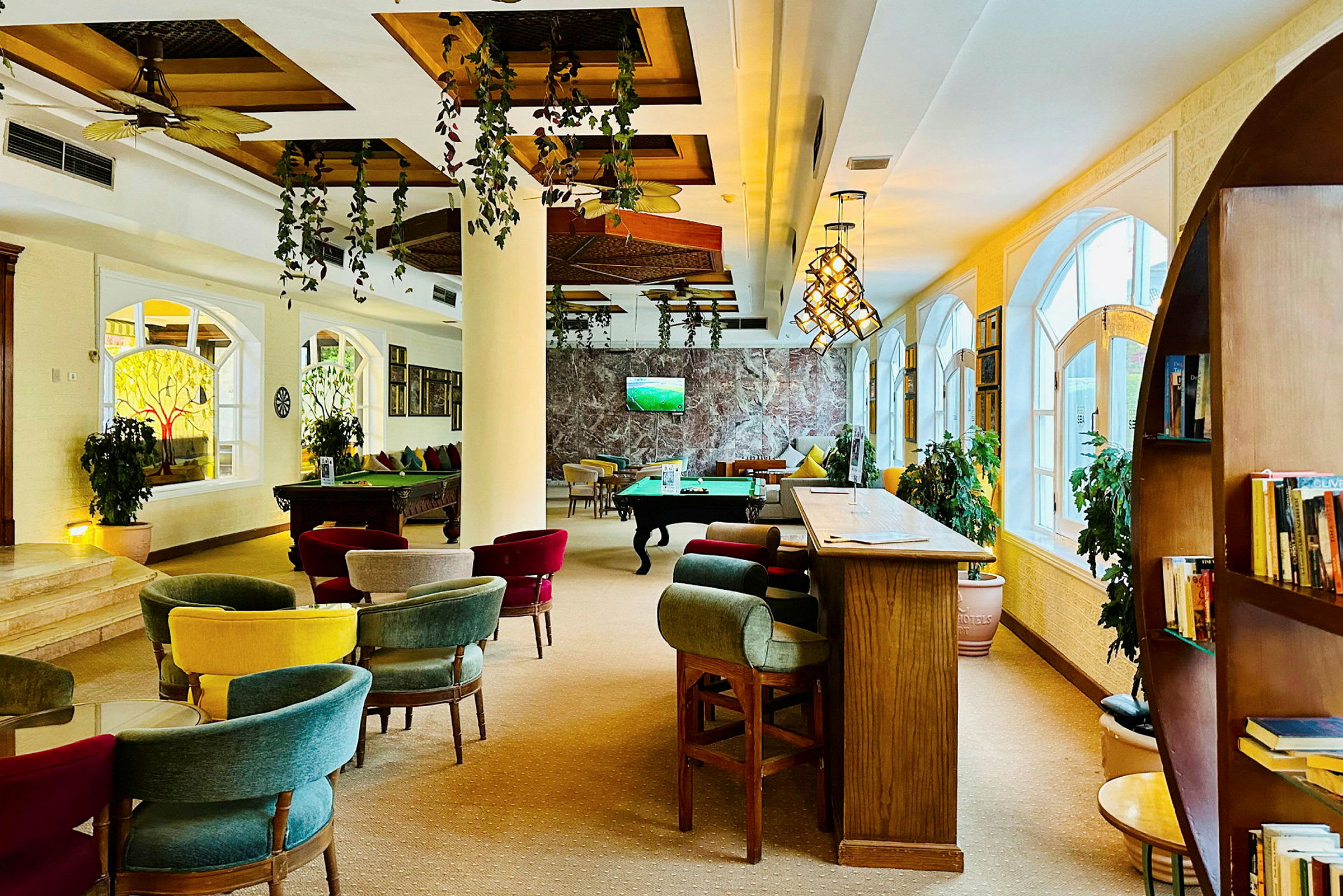Et indendørs område med siddegrupper, et billardbord og en bar med høje siddepladser. Planter og store vinduer dekorerer rummet. (AI-genereret tekst)