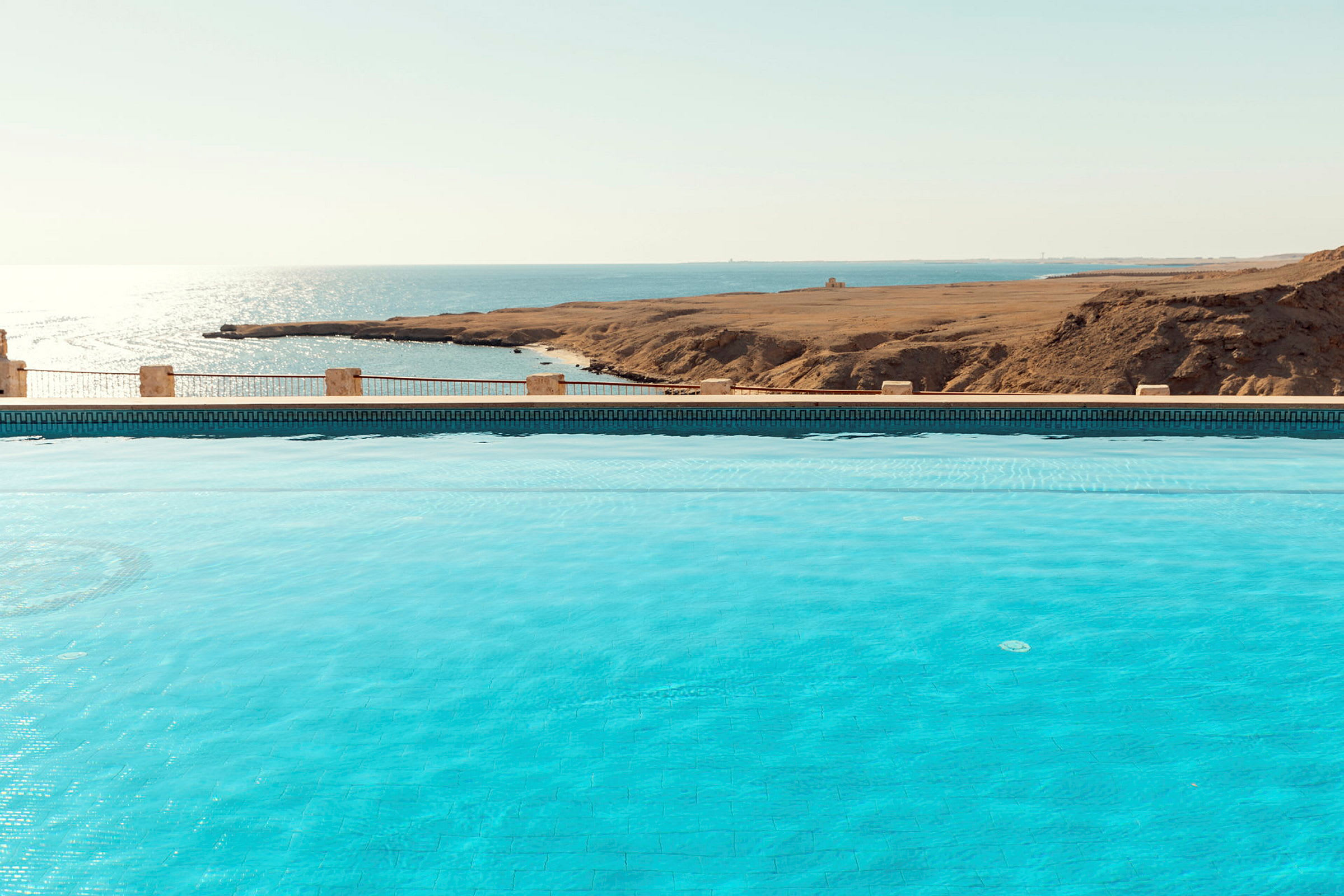 Billede af en pool med klart blåt vand og udsigt over havet og tørt landskab. (AI-genereret tekst)