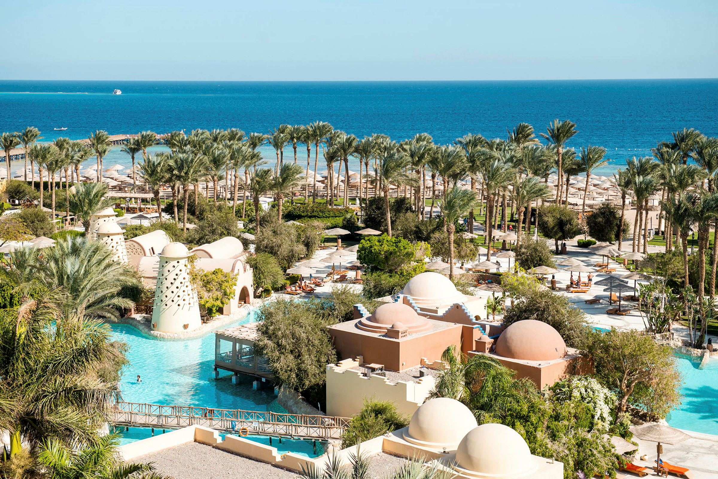 Billede af et hotelområde med swimmingpools, palmer og bygninger med kupler, tæt på en kyst med udsigt til havet. (AI-genereret tekst)