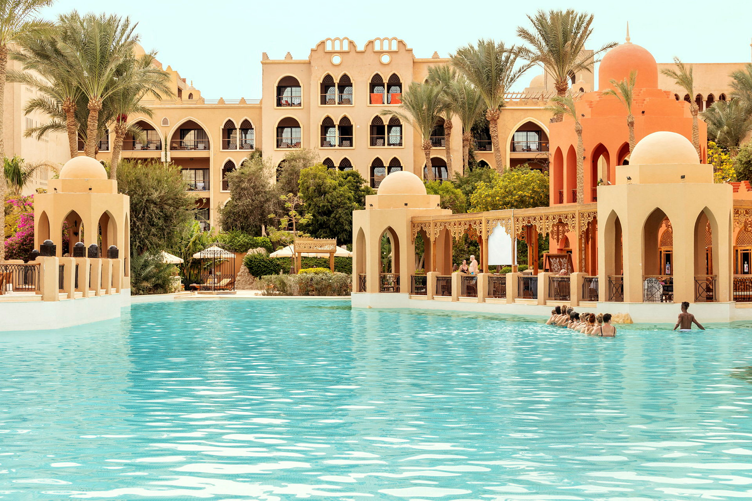 Billedet viser en swimmingpool i forgrunden omgivet af arkitektur i arabisk stil samt palmer og beplantning. (AI-genereret tekst)