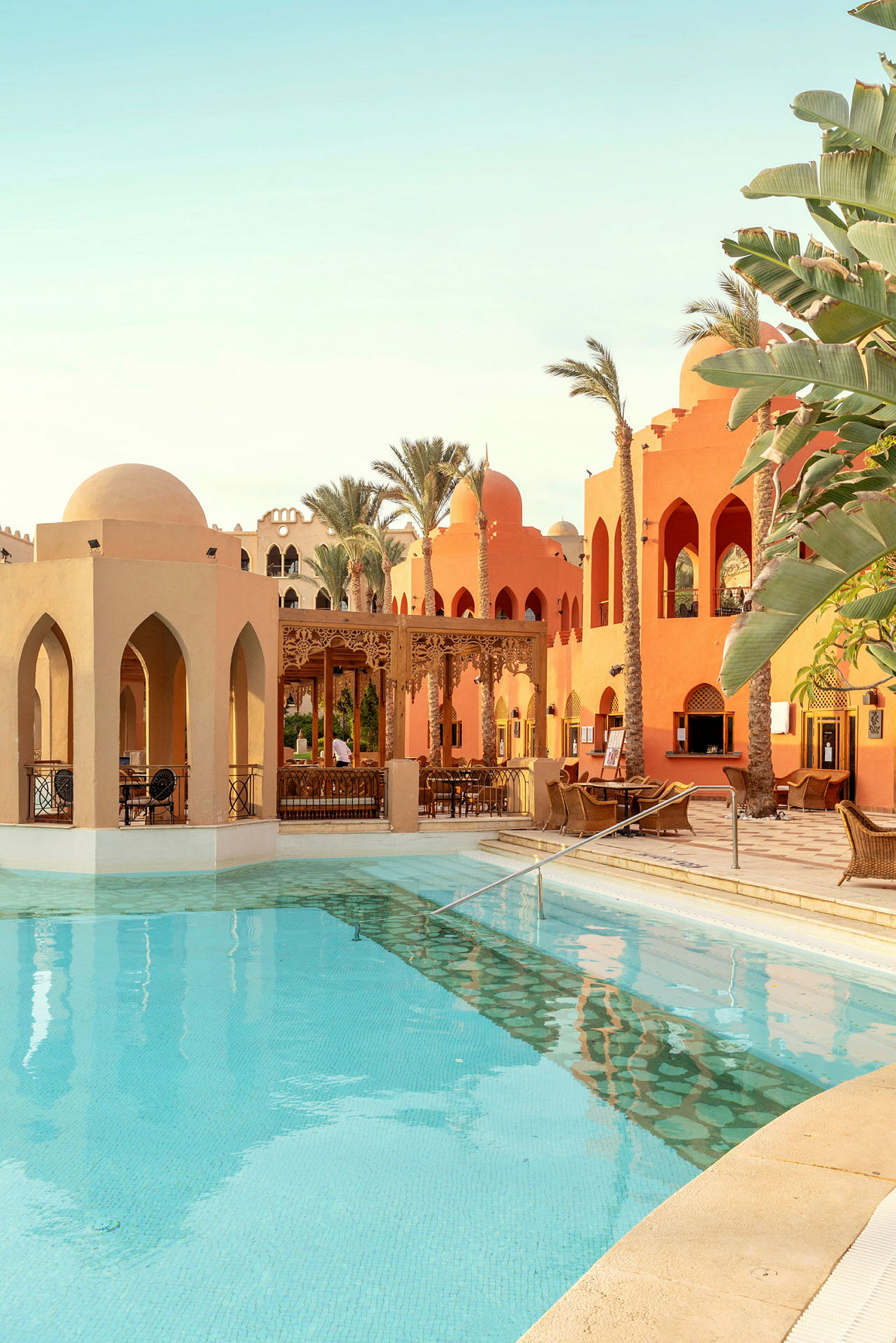 En pool med klart vand omgivet af bygninger med arabisk arkitektur og palmer. (AI-genereret tekst)