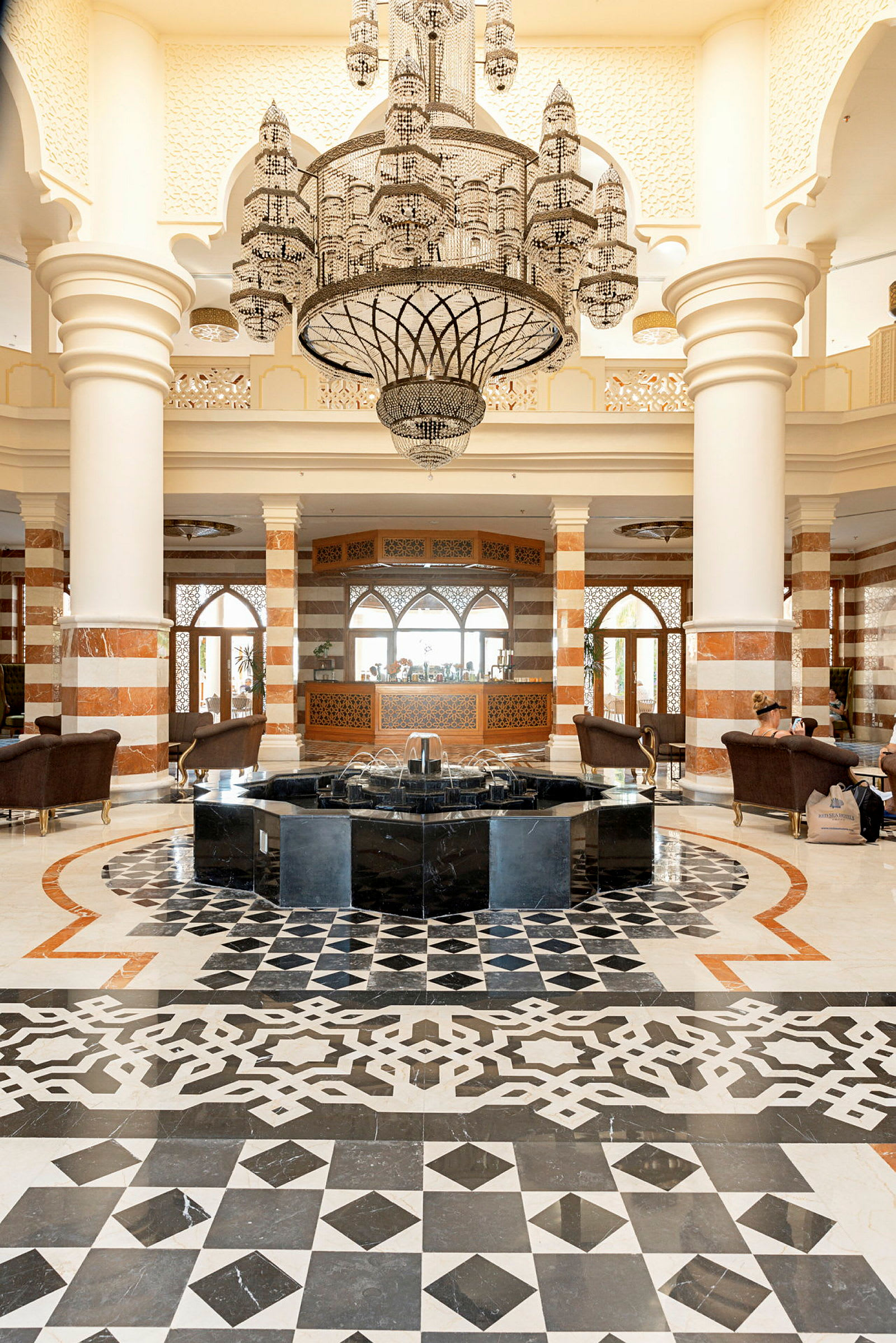Elegant hotellobby med mønstret gulvbelægning, en fontæne i midten og en stor lysekrone hængende ovenover. (AI-genereret tekst)
