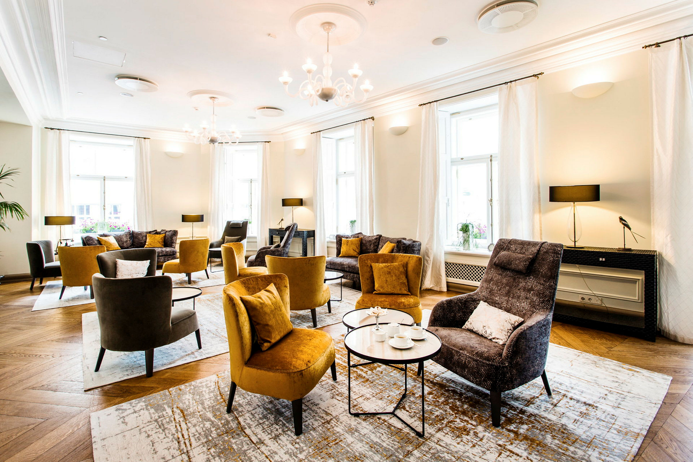 Et elegant indrettet opholdsområde med komfortable stole, sofaer og små sofaborde. Rummet har naturligt lysindfald fra store vinduer og er dekoreret med hvide gardiner. (AI-genereret tekst)