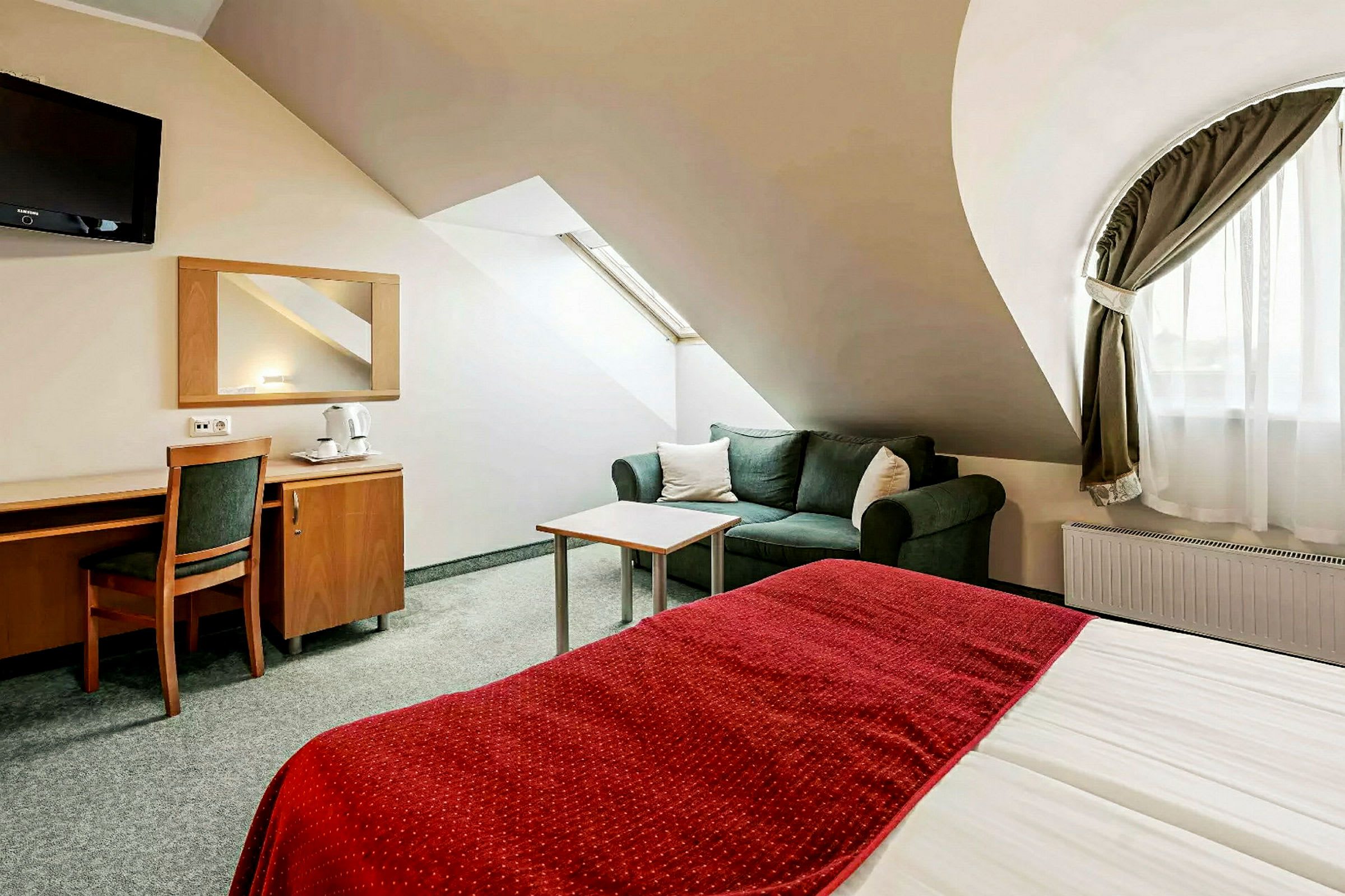 Et hotelværelse med en seng med rødt tæppe, en sofa og et skrivebord med stol. (AI-genereret tekst)