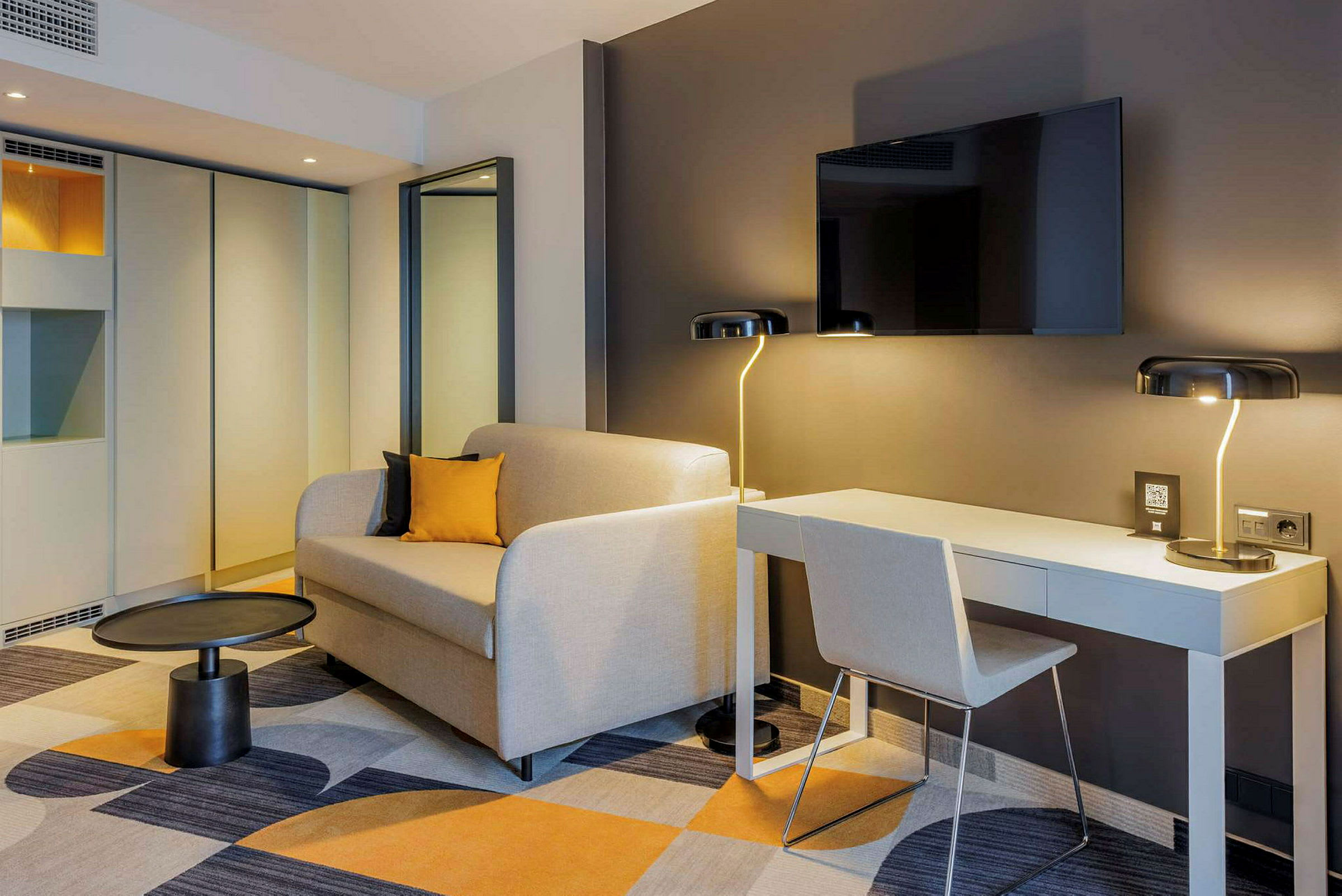 Et moderne hotelværelse med en sofa, et skrivebord og en vægmonteret TV. (AI-genereret tekst)