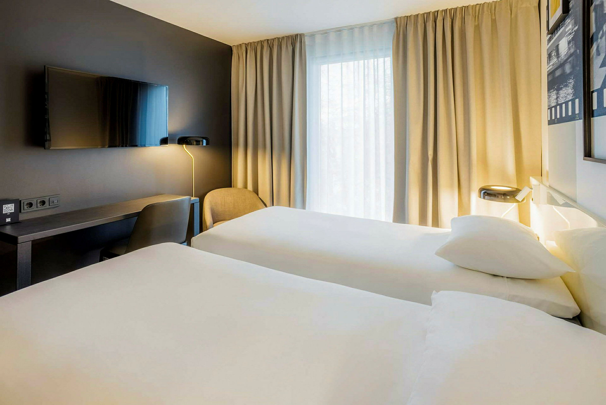 Hotelværelse med to enkeltsenge, et skrivebord, en stol og et vægmonteret TV samt gardiner ved et vindue. (AI-genereret tekst)