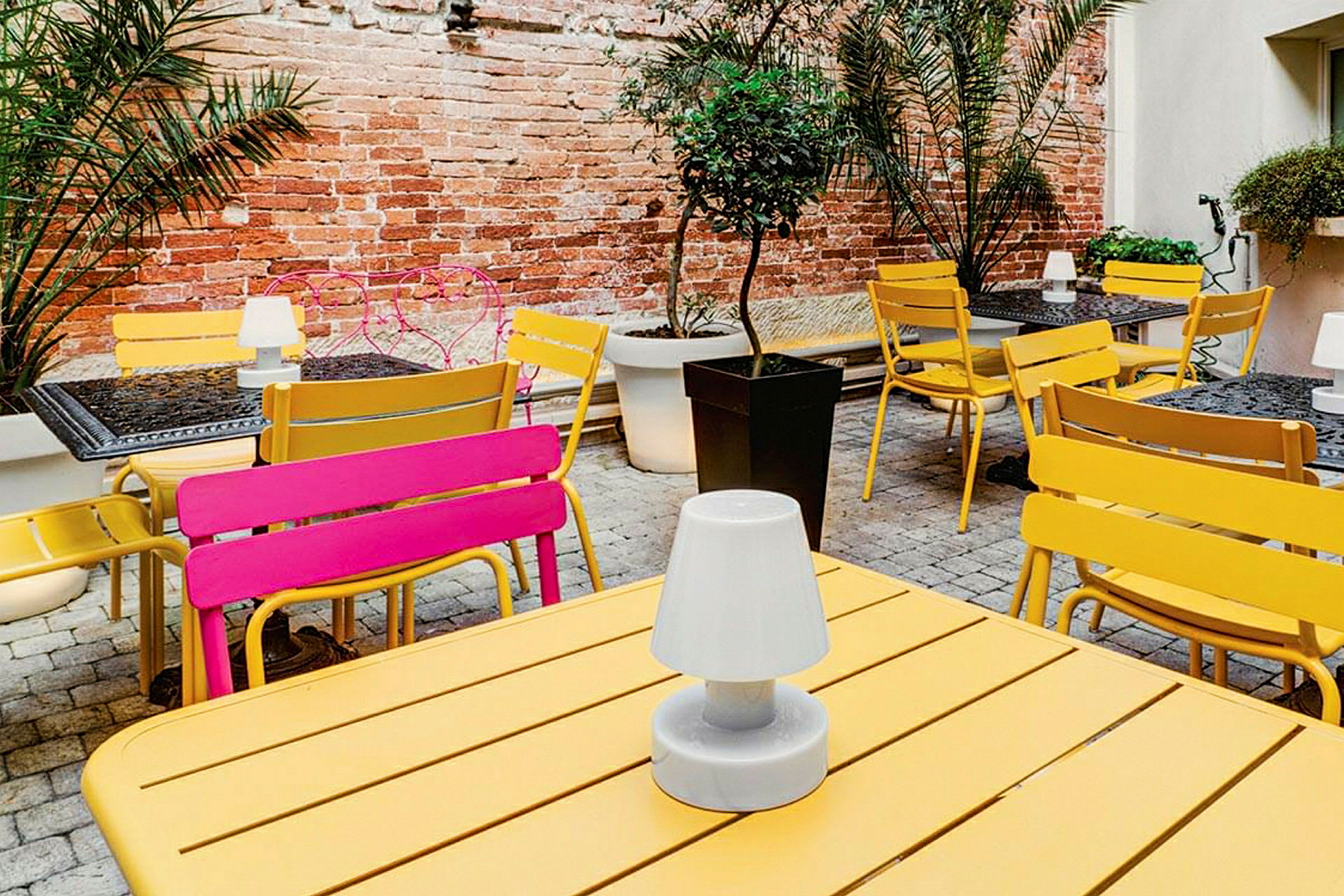 Flere borde og stole er placeret på en udendørs terrasse med planter og en murstensvæg. (AI-genereret tekst)