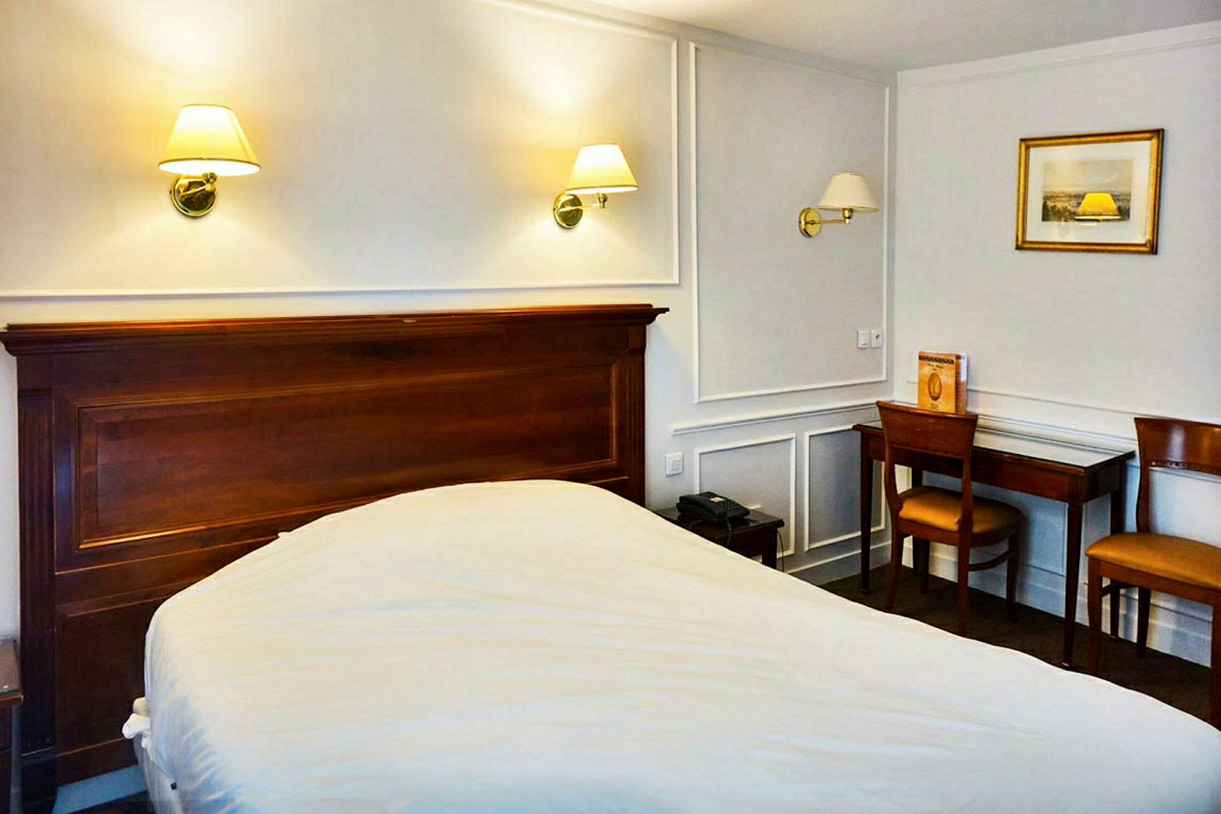 Et hotelværelse med en seng, træbord og stole samt væglamper over sengen. (AI-genereret tekst)