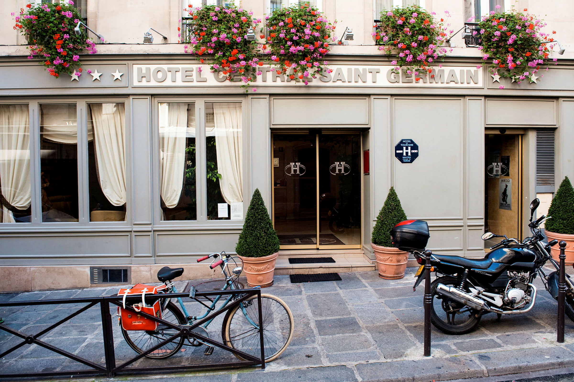 Eksteriøret af et hotel med skiltet 'Hotel Central Saint Germain' med blomsterkrukker over indgangen samt en cykel og motorcykel parkeret foran. (AI-genereret tekst)