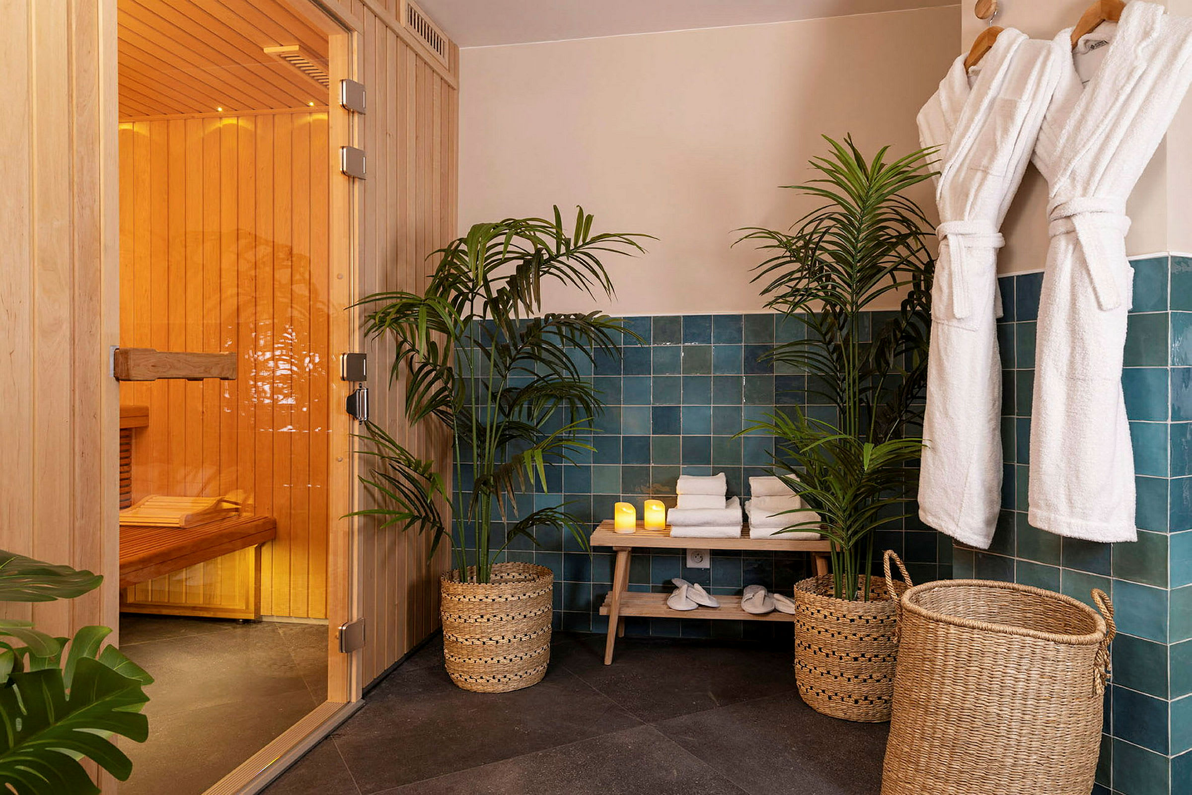Et spaområde med en sauna, grønne planter, foldede håndklæder på en bænk, nogle levende lys og hængende hvide badekåber. (AI-genereret tekst)