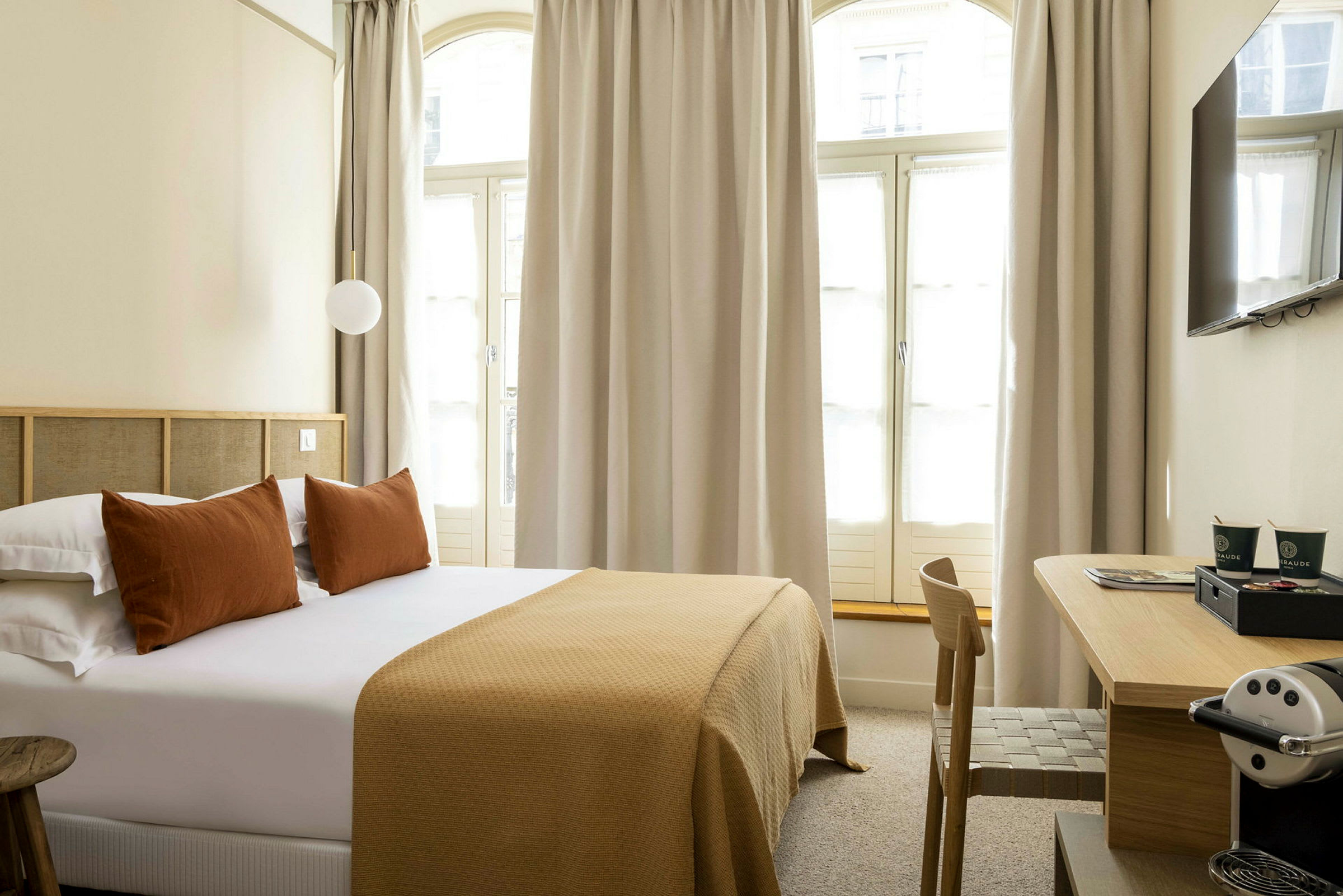 Hotelværelse med en dobbeltseng, puder og et sengetæppe i brune nuancer samt et skrivebord med en kaffemaskine. (AI-genereret tekst)