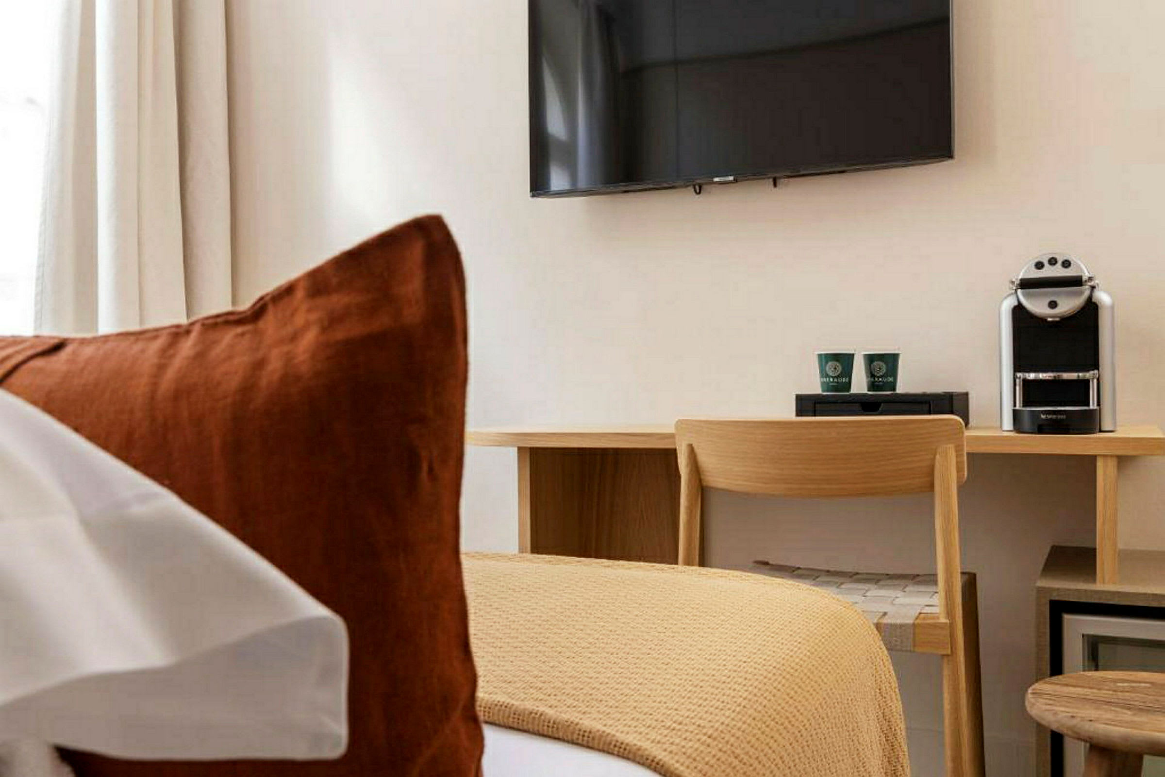 Et hotelværelse med en seng i forgrunden, en skrivebordsplads med stol, kaffemaskine og en vægmonteret TV. (AI-genereret tekst)