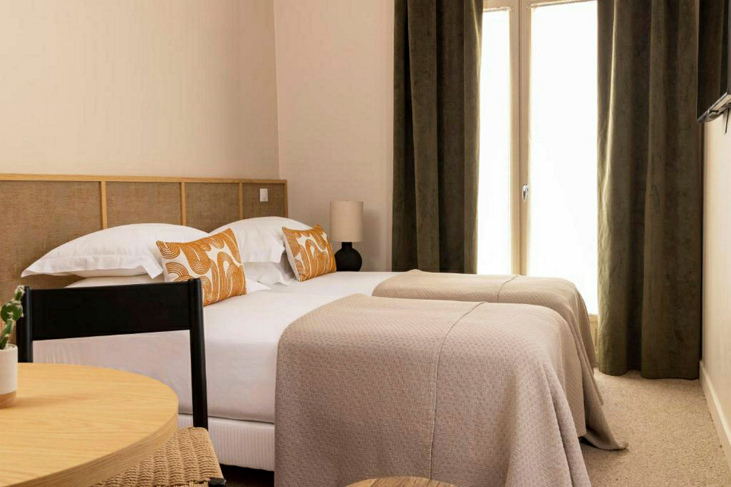 Et hotelværelse med to enkeltsenge dækket med beige sengetæpper og dekoreret med orange mønstrede puder. (AI-genereret tekst)