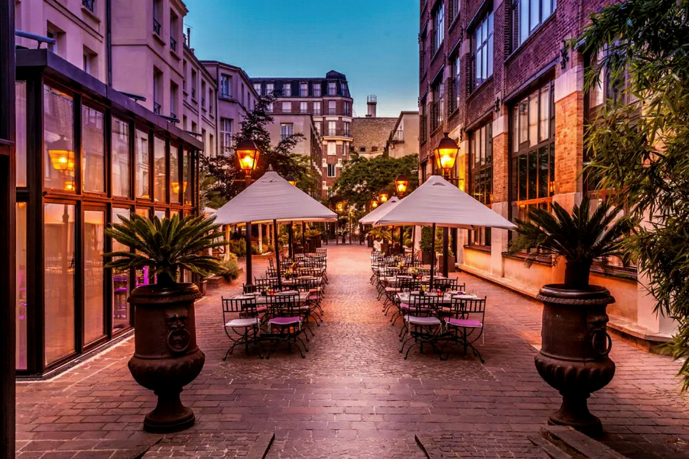 Et udendørs restaurantområde med borde og parasoller omgivet af bygninger. (AI-genereret tekst)