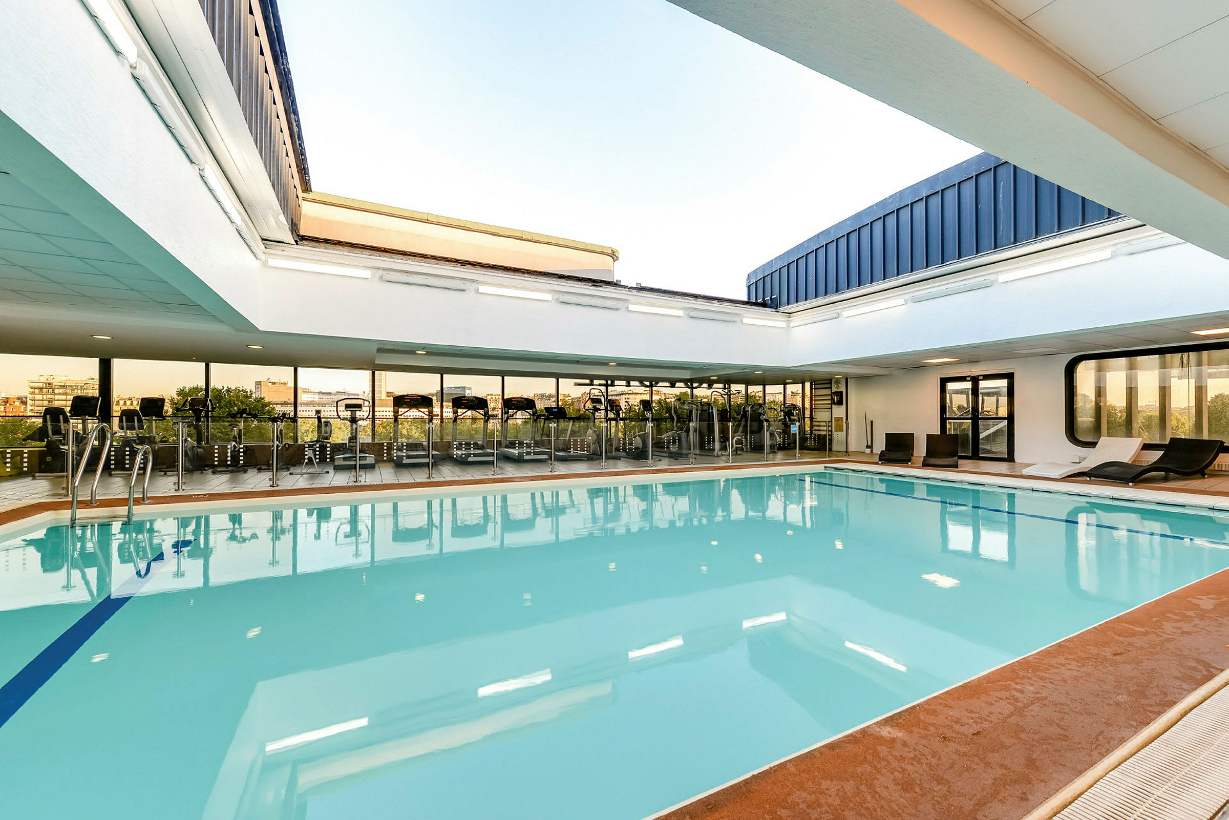 En indendørs swimmingpool med omkringliggende liggestole og store vinduer mod et træningscenter. (AI-genereret tekst)