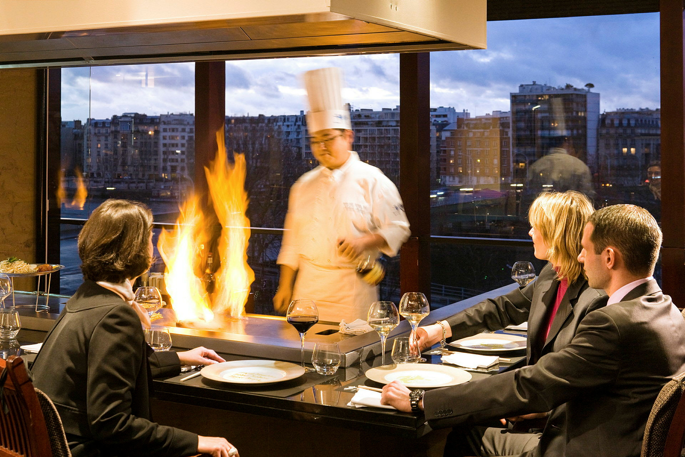 En gruppe mennesker sidder ved et bord og nyder et måltid, mens kokken tilbereder mad med flammeeffekt i en restaurant med store vinduer og aftenudsigt. (AI-genereret tekst)