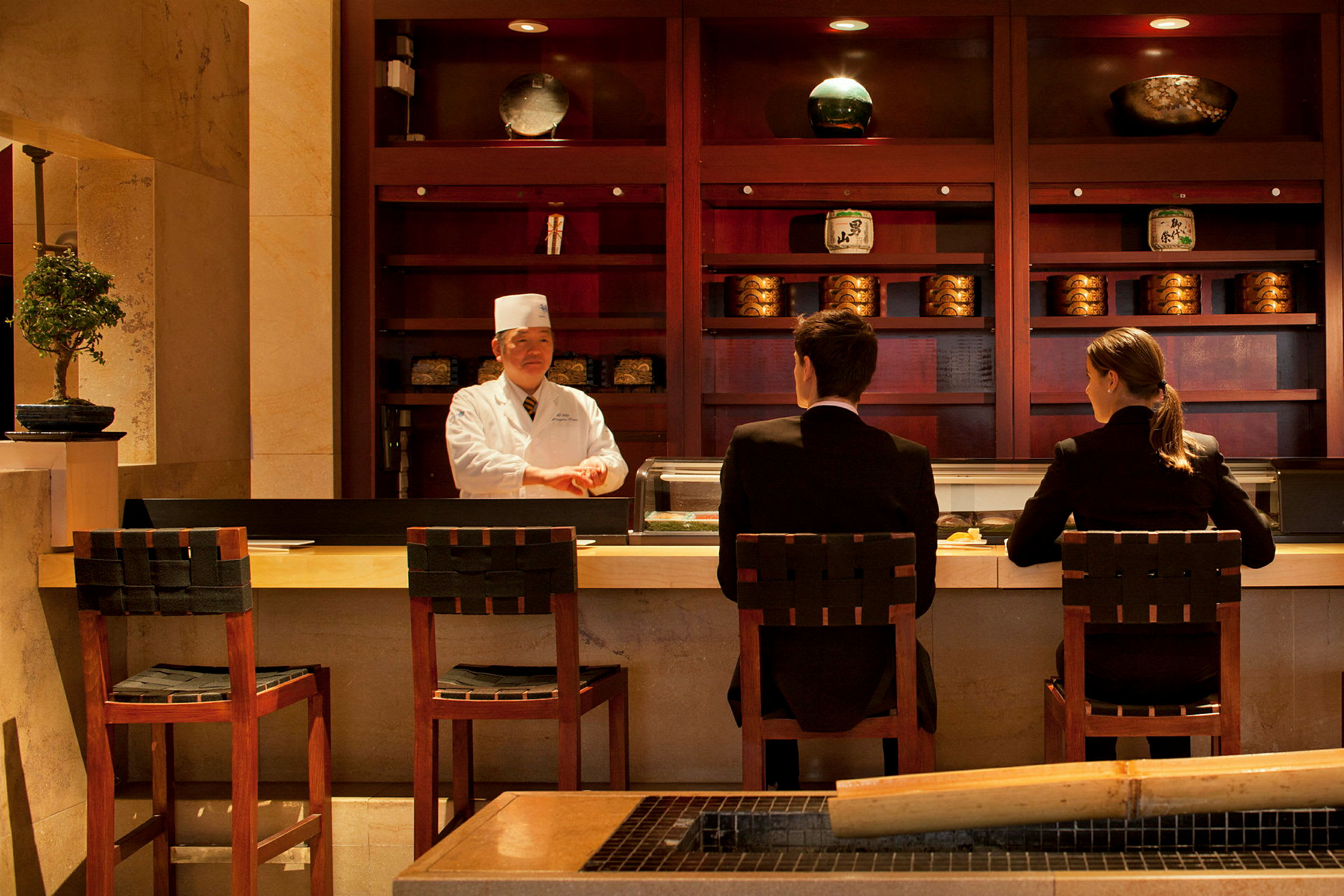 To personer sidder ved en sushi-bar med en kok bag disken. (AI-genereret tekst)