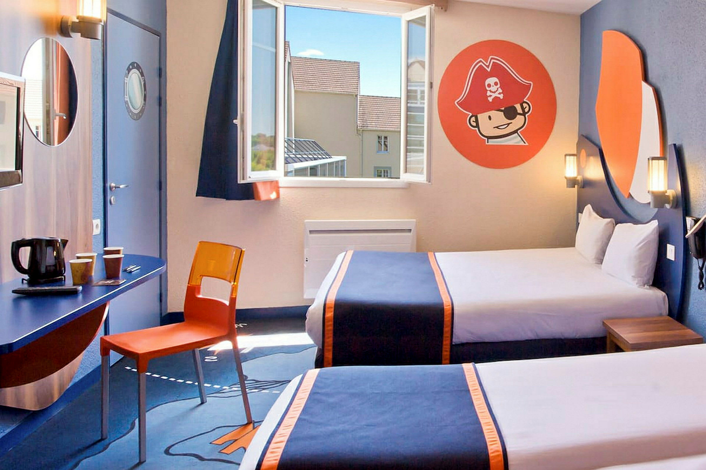 Et hotelværelse med to senge forsynet med mørkeblå sengetæpper og orange detaljer. Der er et vindue med åben udsigt, en væg med pirattema på dekorative vægmalerier samt et skrivebord med en orange stol og kaffemaskine under et rundt spejl. (AI-genereret tekst)