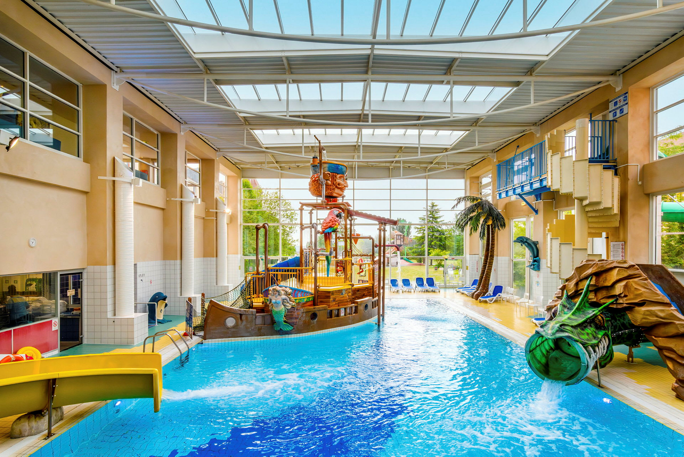 En indendørs pool med et piratskib og vandrutsjebaner, omgivet af liggestole og planter. (AI-genereret tekst)