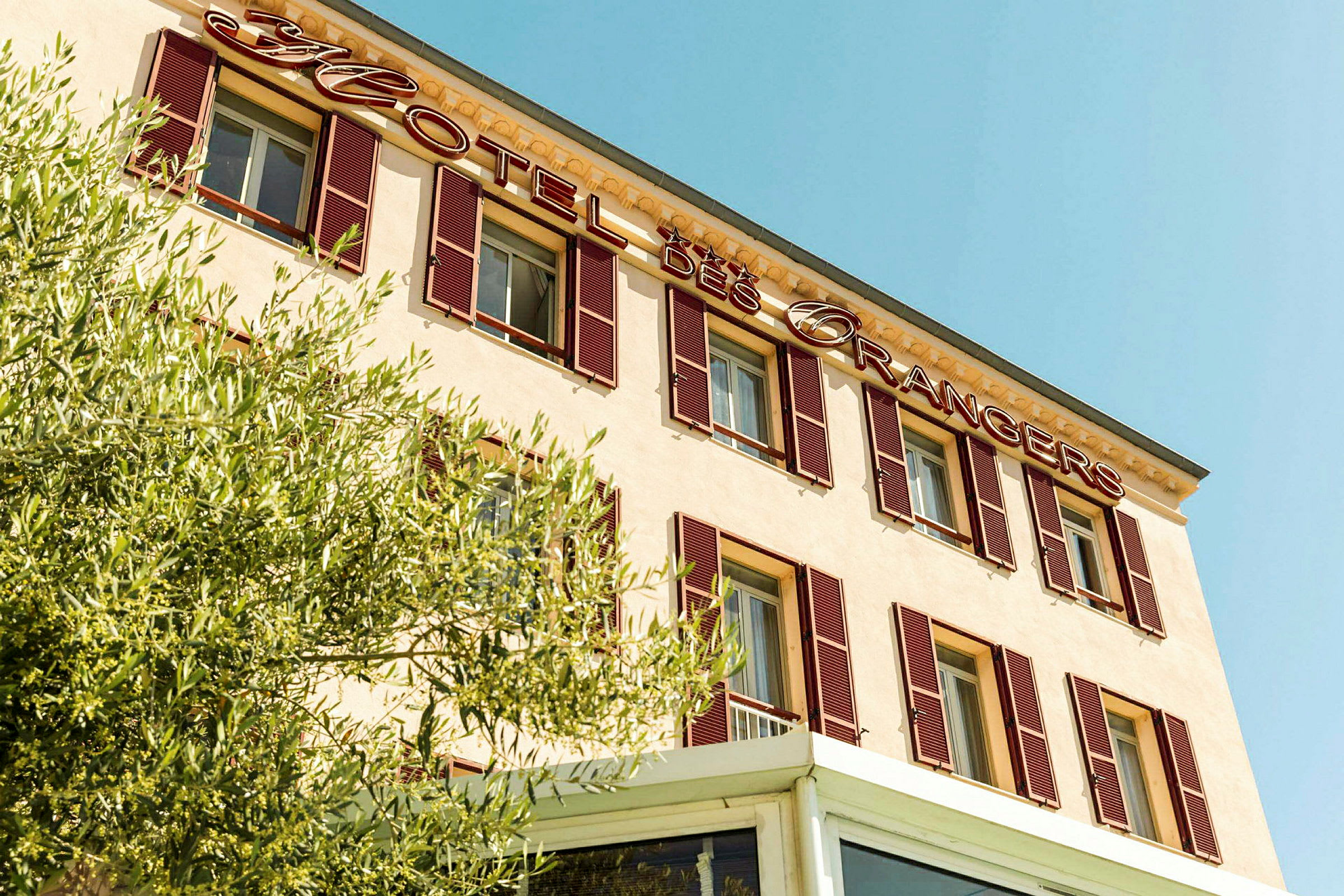 Facaden af en hotelbygning med skiltet 'Hotel des Orangers' og vegetation i forgrunden. (AI-genereret tekst)
