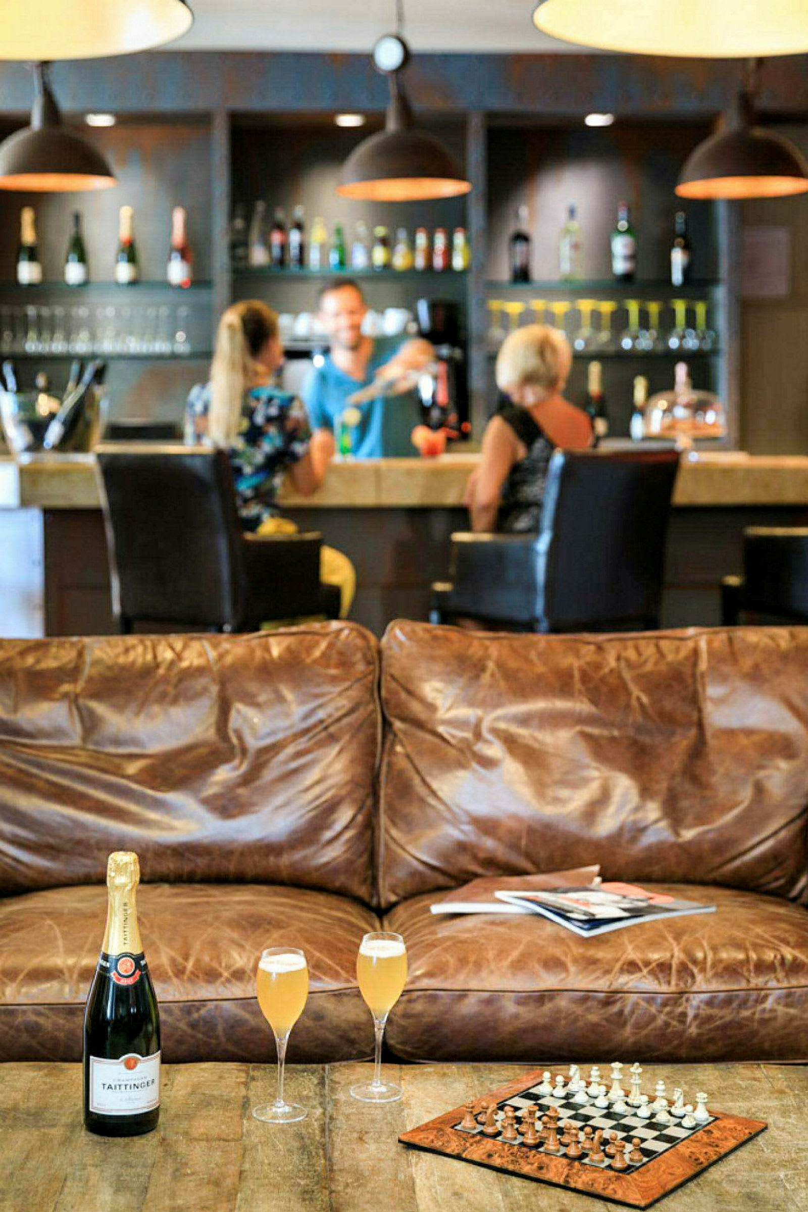 Interiørbillede af en bar med flasker i baggrunden, en lædersofa, et skakspil og et bord med champagneglas i forgrunden. (AI-genereret tekst)