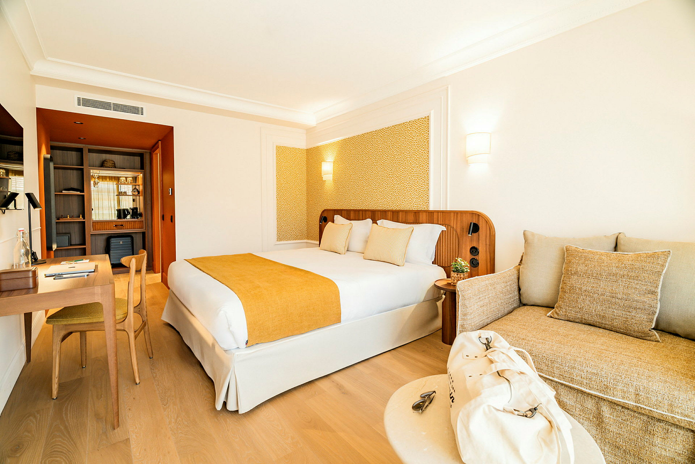 Et hotelværelseinteriør med en dobbeltseng med gult sengetæppe og puder, et skrivebord med stol til venstre og en sofa med beige puder til højre. (AI-genereret tekst)