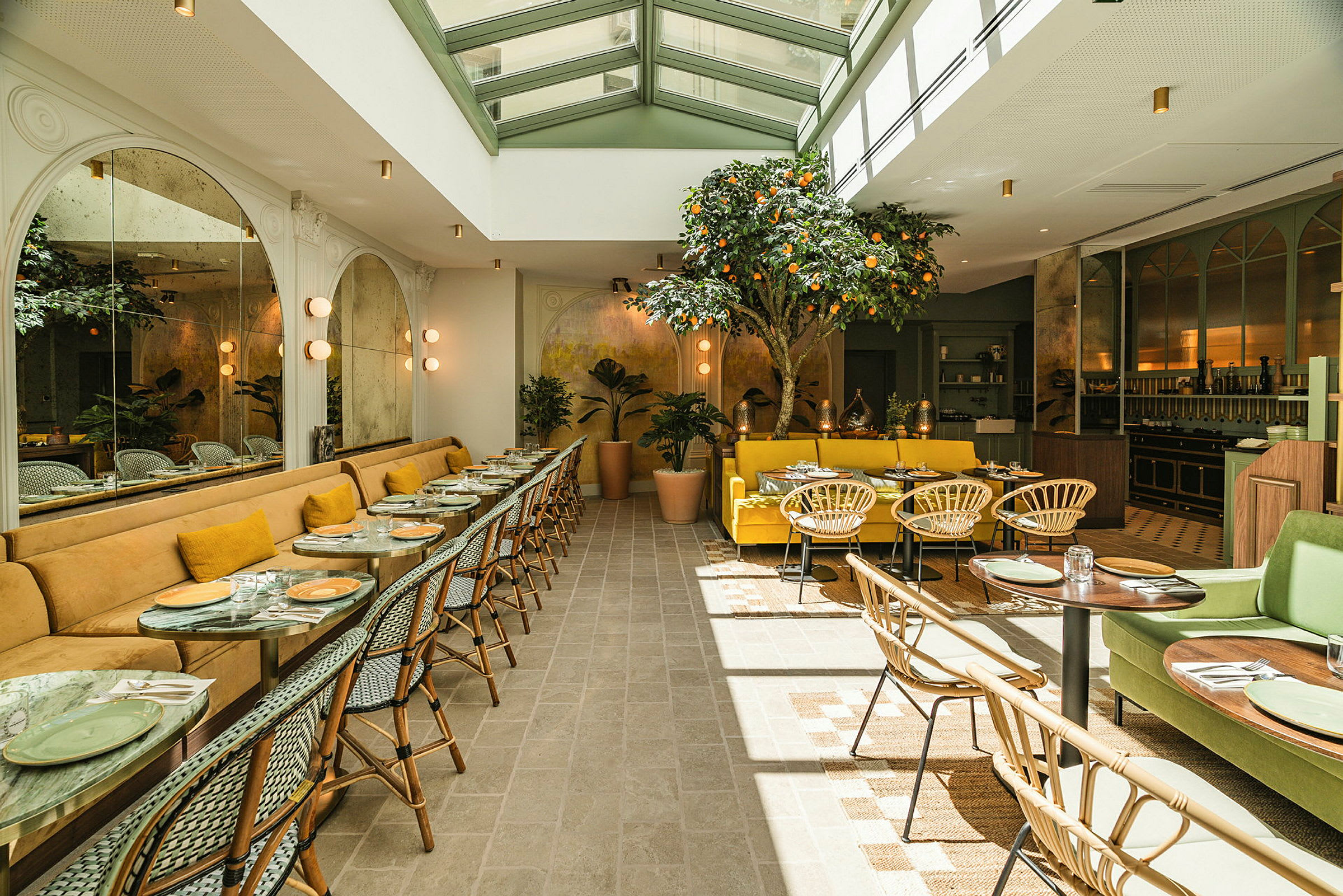 En lys og elegant restaurant med siddepladser ved borde og sofaer. Dekorationer inkluderer et citrus træ og grønne planter. (AI-genereret tekst)