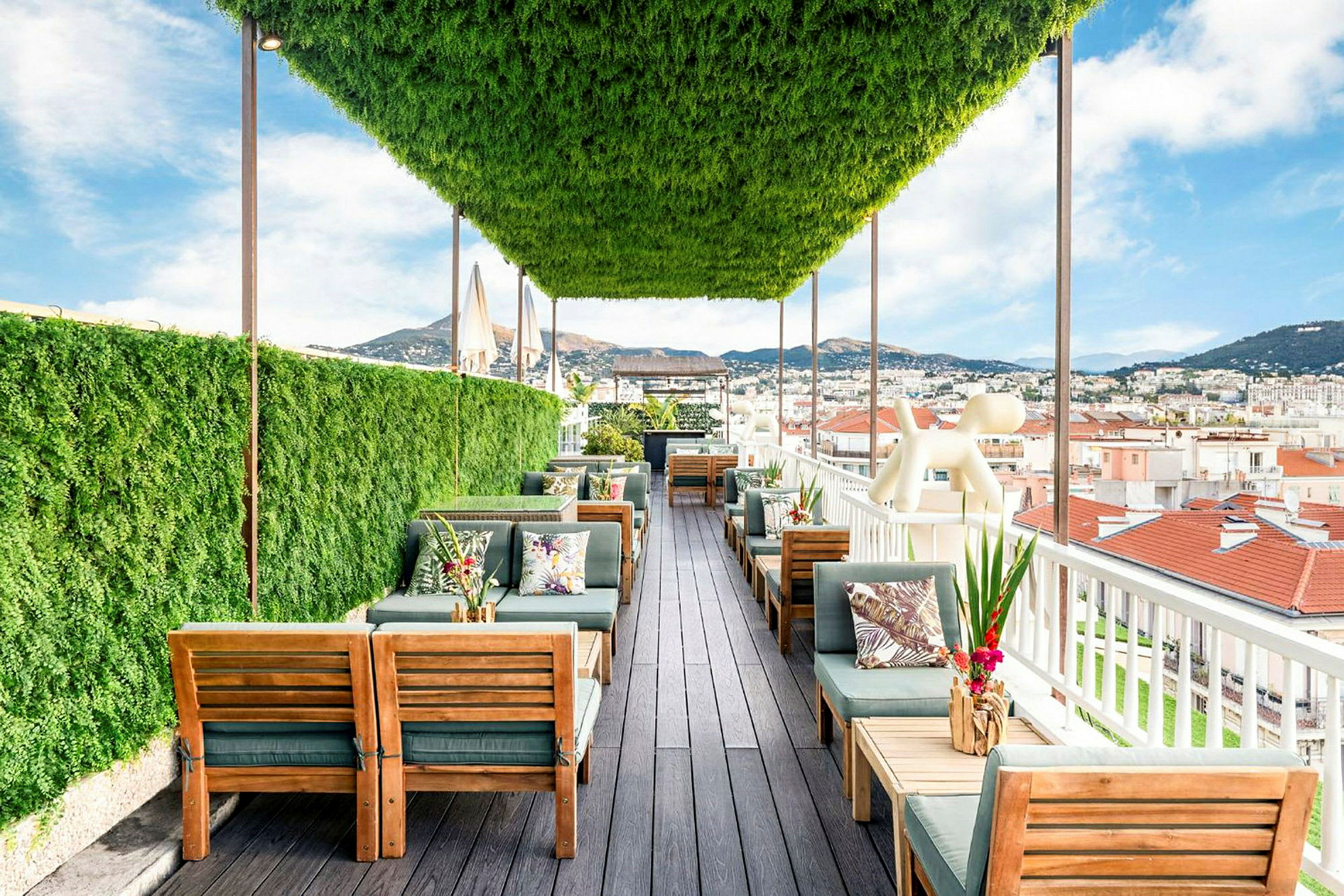 En tagterrasse med grønt tag og planter, møbleret med træmøbler og borde, med udsigt over hustage og bjerge. (AI-genereret tekst)