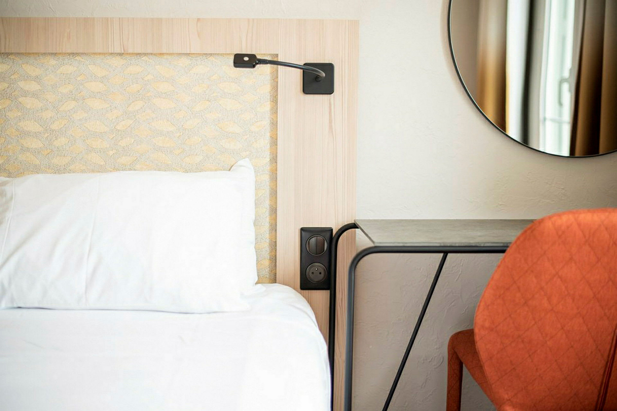 En hotelseng med en hvid pude og et natbord ved siden af, samt en orange stol foran et bord og et rundt spejl på væggen. (AI-genereret tekst)