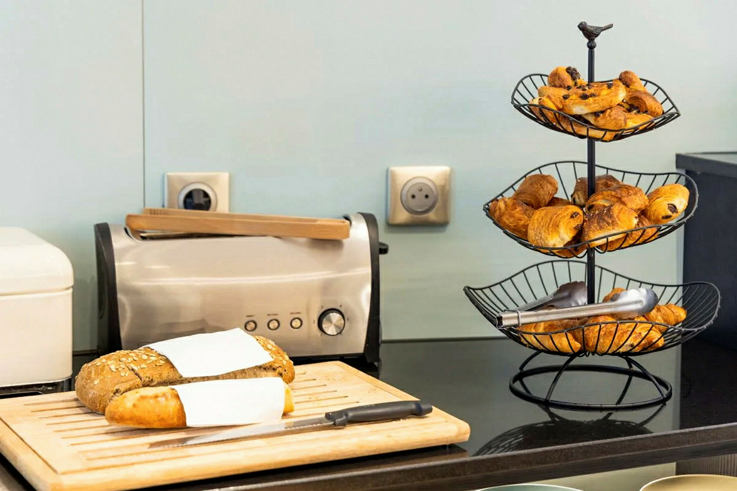 Billede af et skærebræt med en brødrister og en brødstativ med croissanter på en køkkenbord. (AI-genereret tekst)