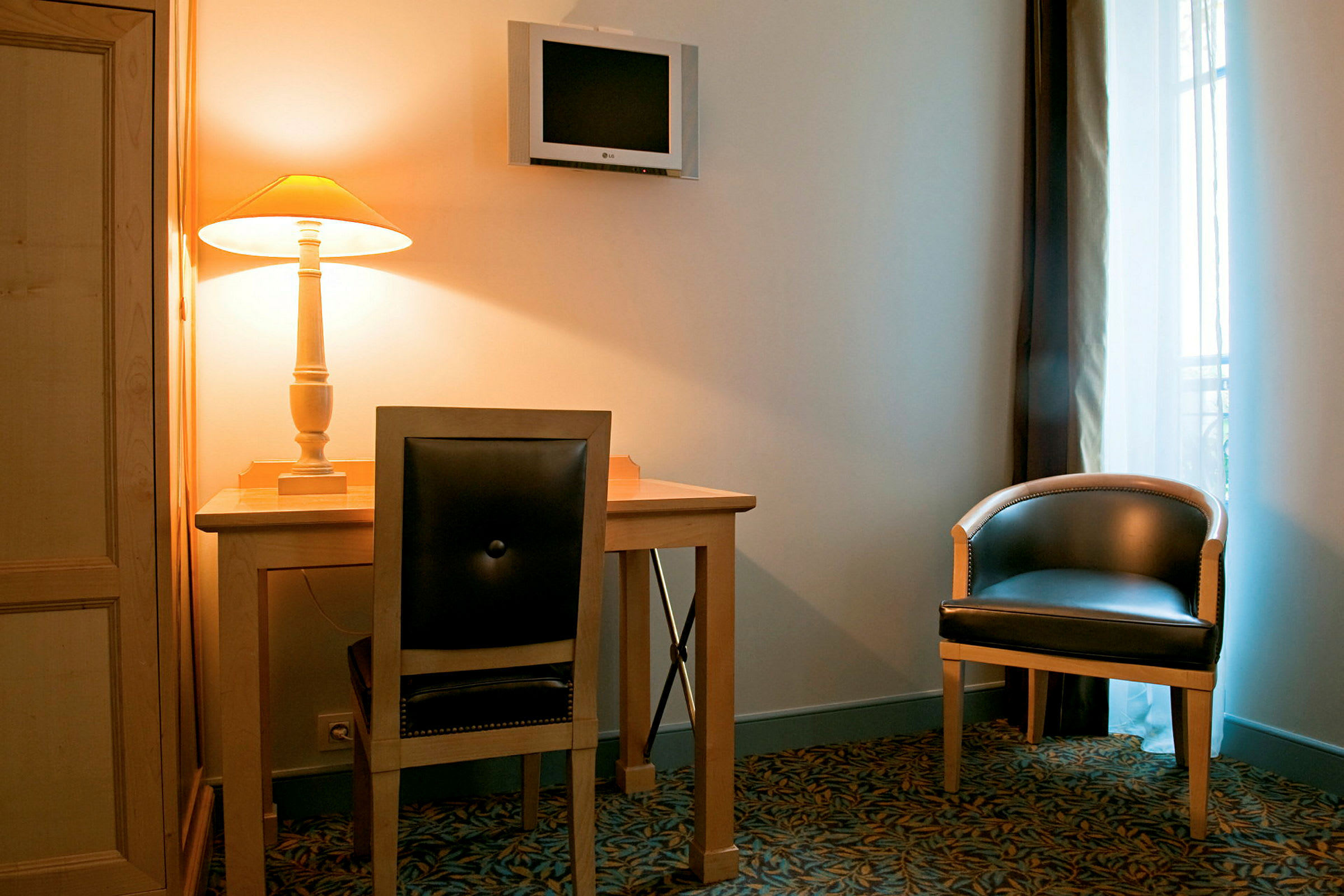 Et interiør med et skrivebord og stol, et vægmonteret fjernsyn og en lænestol ved siden af et vindue. Der står en tændt bordlampe på skrivebordet. (AI-genereret tekst)