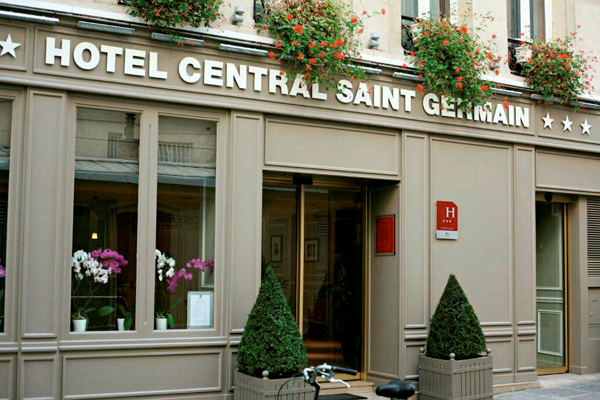Billede af hotelindgangen til Hotel Central Saint Germain med blomsterdekorationer og skiltet synligt. (AI-genereret tekst)