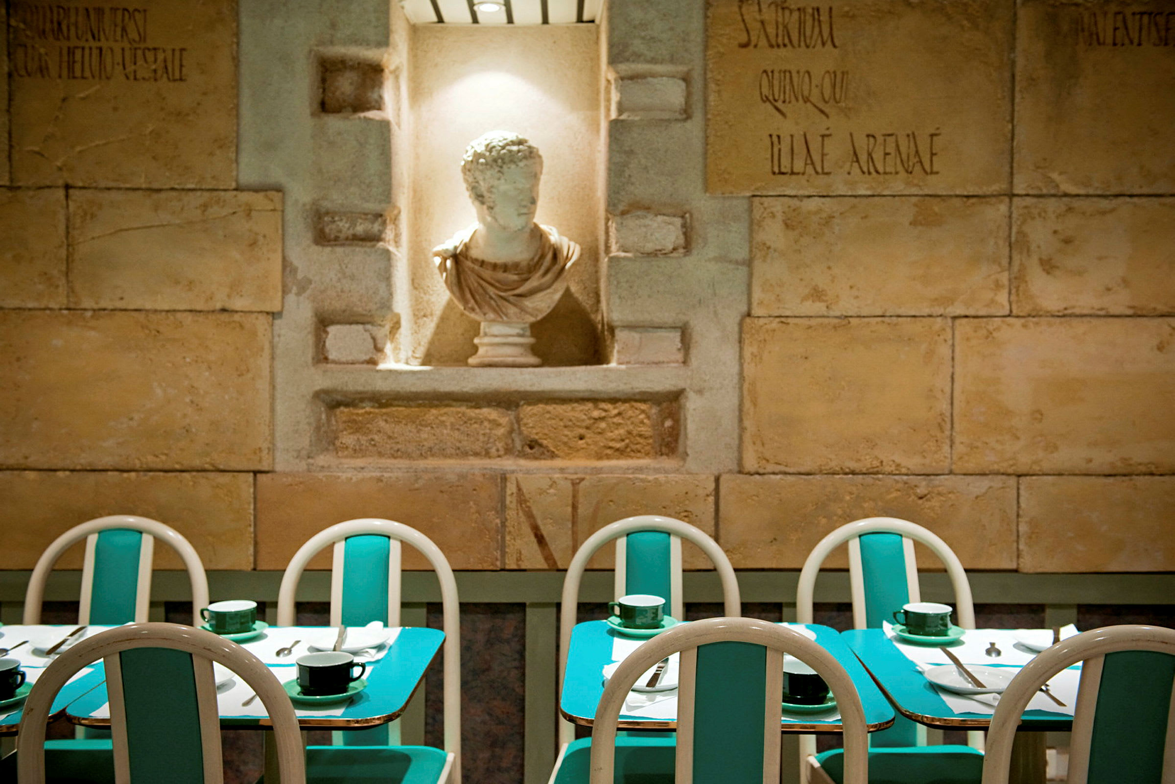 Billedet viser en restaurant med turkise borde og stole samt en hvid skulptur i en niche på væggen. (AI-genereret tekst)