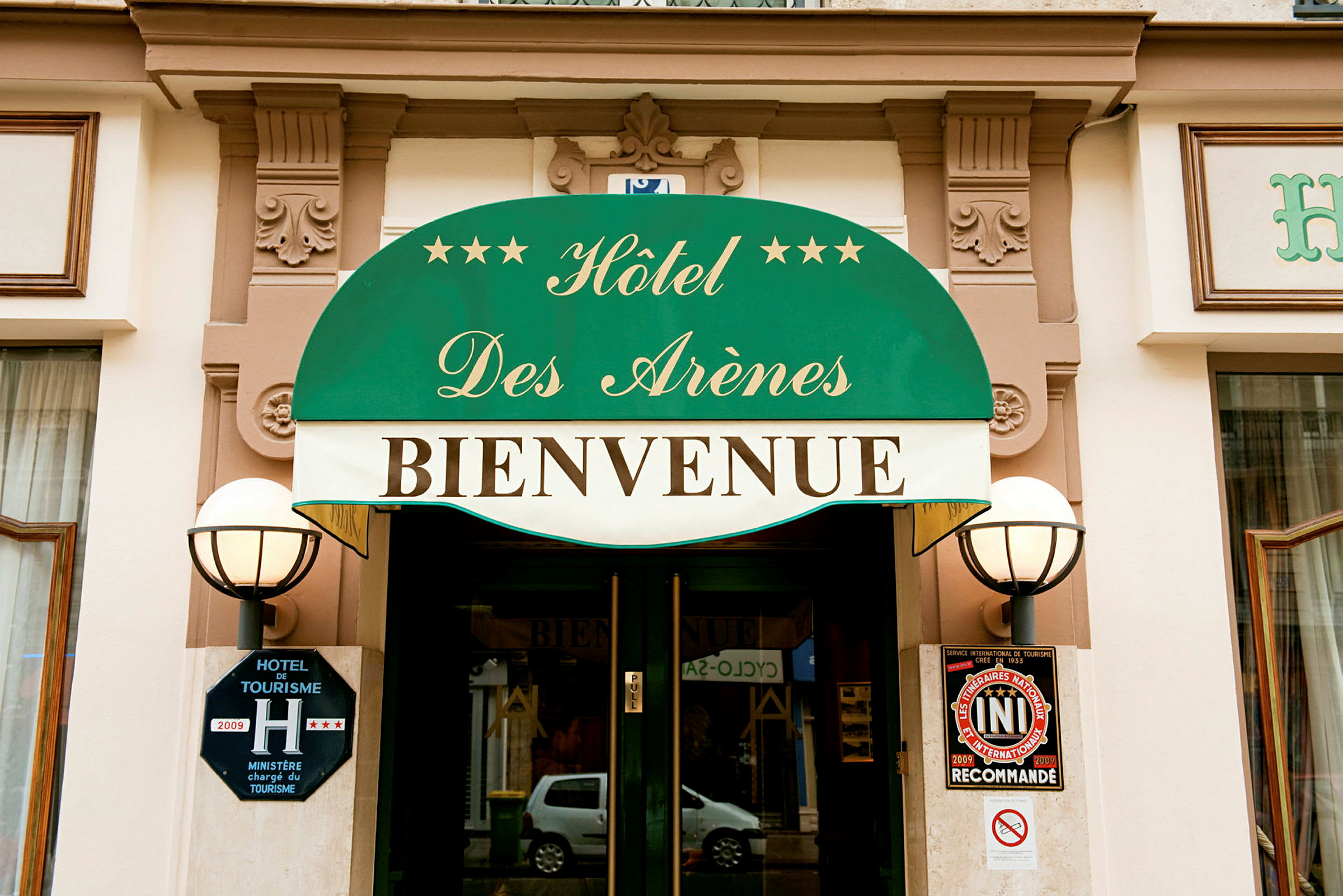 Indgangen til Hôtel Des Arènes med grøn markise og teksten 'Bienvenue'. (AI-genereret tekst)