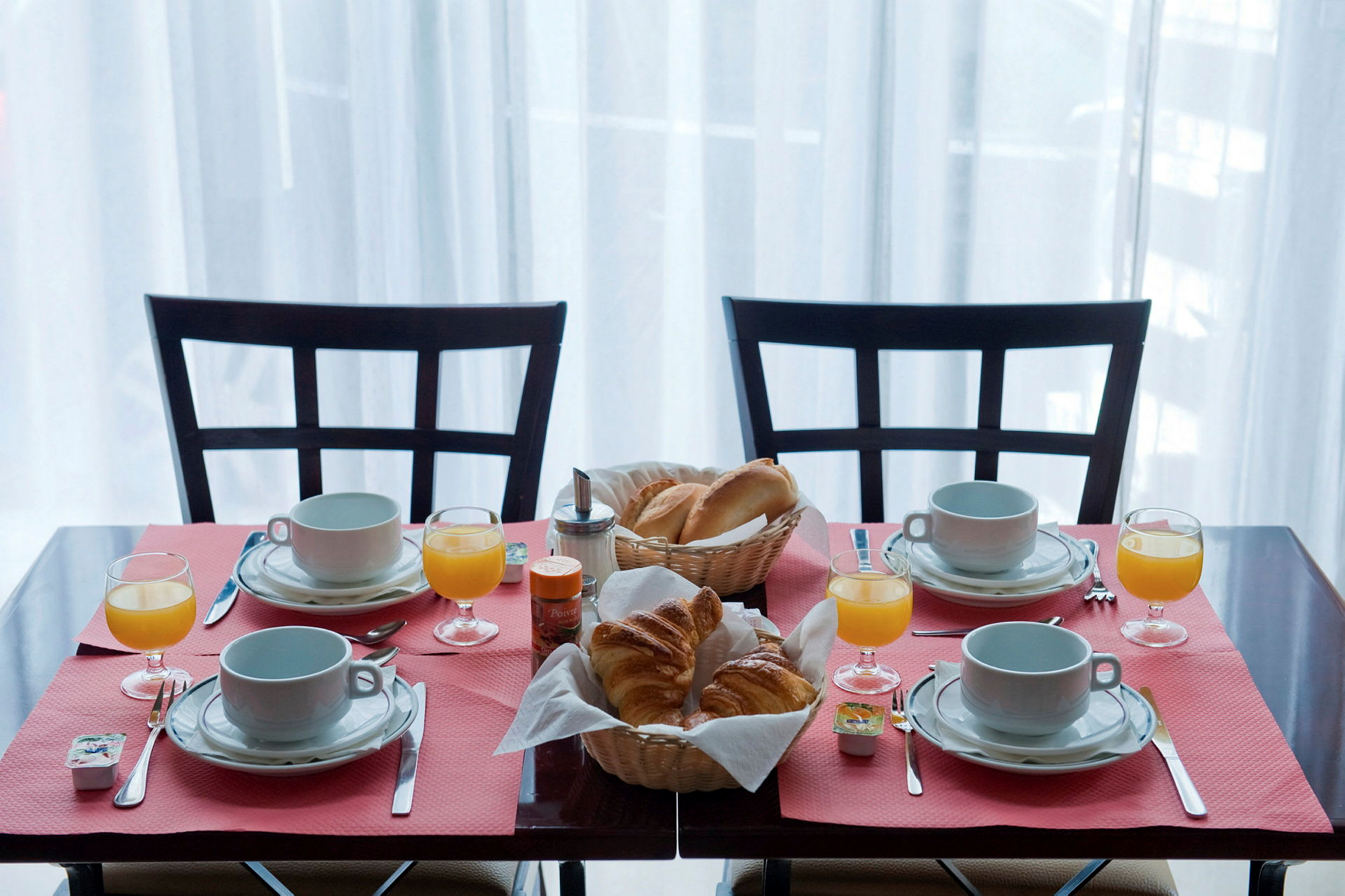 Et morgenbord dækket til fire personer med kaffekopper, glas med appelsinjuice og kurve med brød og croissanter. (AI-genereret tekst)