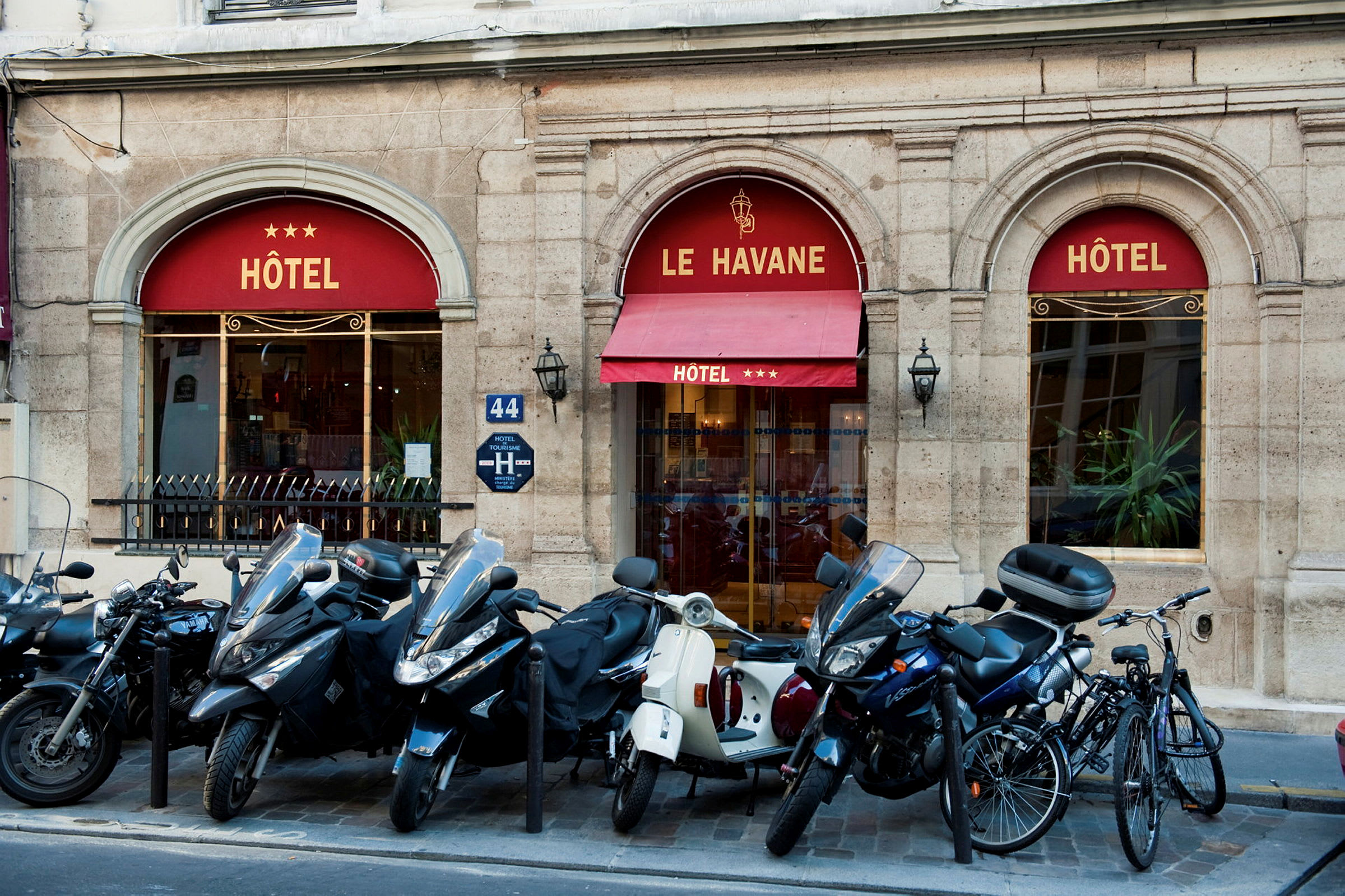 Billede, der viser indgangen til et hotel med navnet Le Havane, tre stjerner vises på markisen. Foran bygningen er der parkeret scootere og cykler. (AI-genereret tekst)