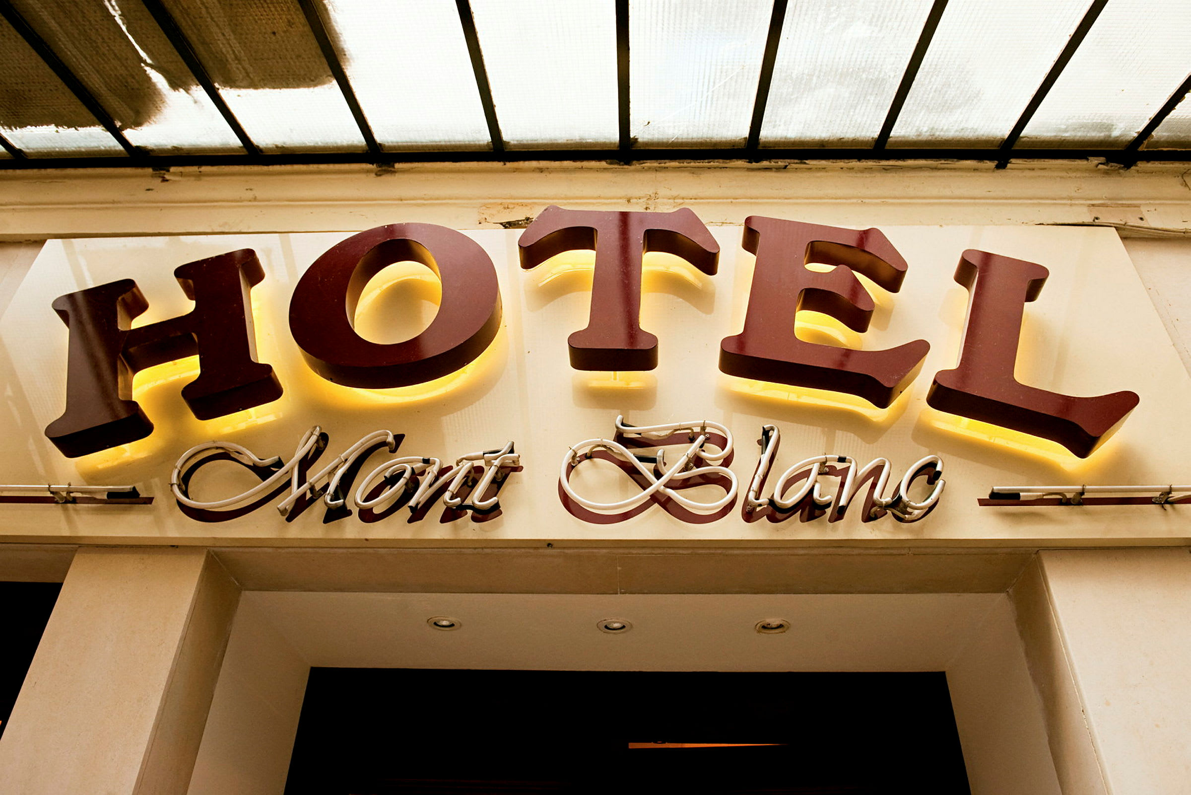 Et hotelskilt med teksten "HOTEL Mont Blanc", hvor "HOTEL" er i store oplyste bogstaver, og "Mont Blanc" er i en kursiv dekorativ stil. (AI-genereret tekst)