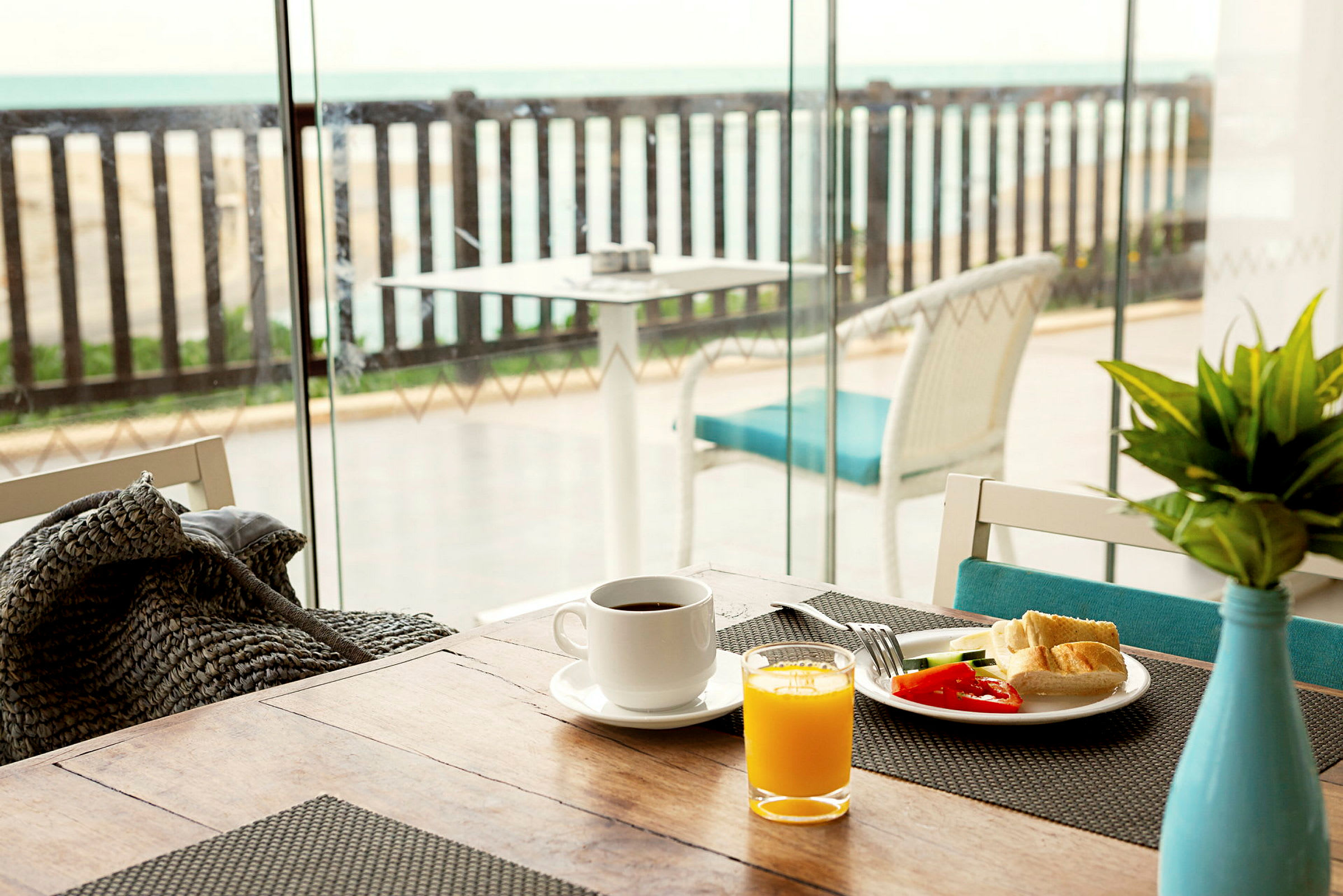 Morgenmadsbord med kaffe, juice og grøntsager nær et glasvindue med udsigt over en terrasse med havet i baggrunden. (AI-genereret tekst)