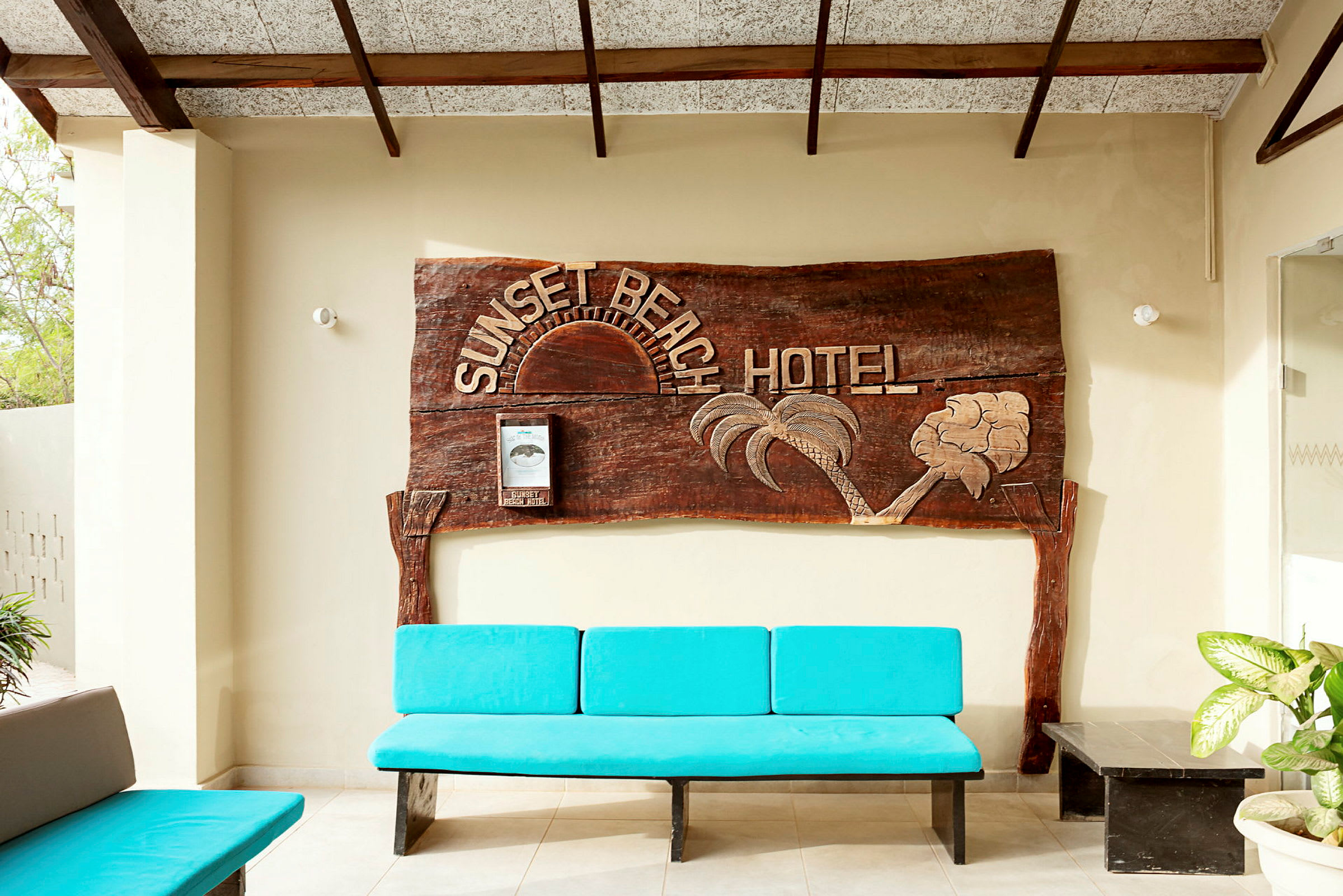 Et træskilt med teksten "Sunset Beach Hotel" pryder væggen. Foran står en blå bænk og nogle planter. (AI-genereret tekst)