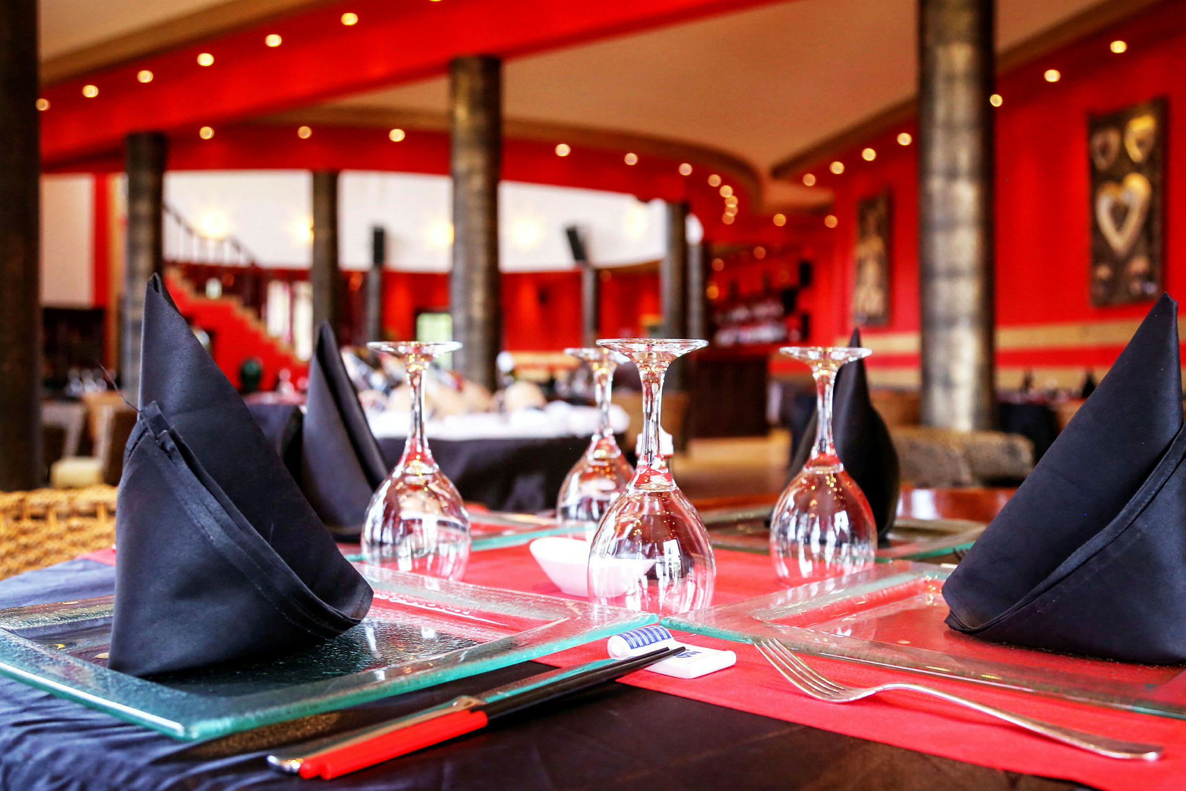 Nærbillede af borddækning på en restaurant med glas og sorte stofservietter på et bord, omgivet af stilfulde røde vægge. (AI-genereret tekst)