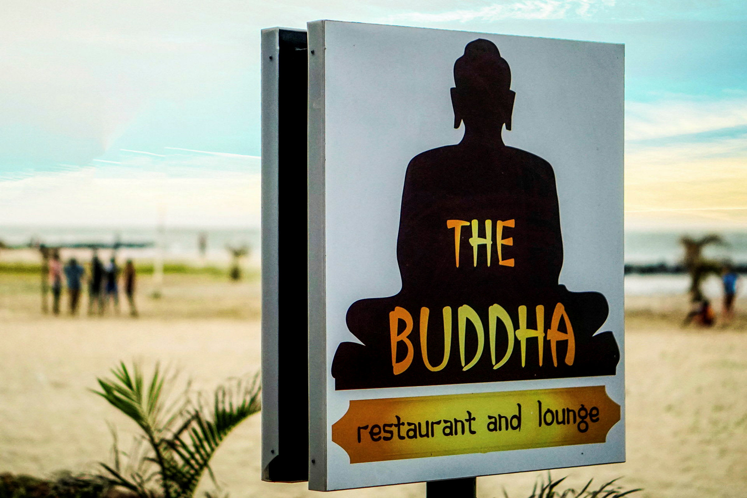 Et skilt med teksten 'The Buddha restaurant and lounge' med en silhuet af en siddende Buddha foran en strandbaggrund. (AI-genereret tekst)