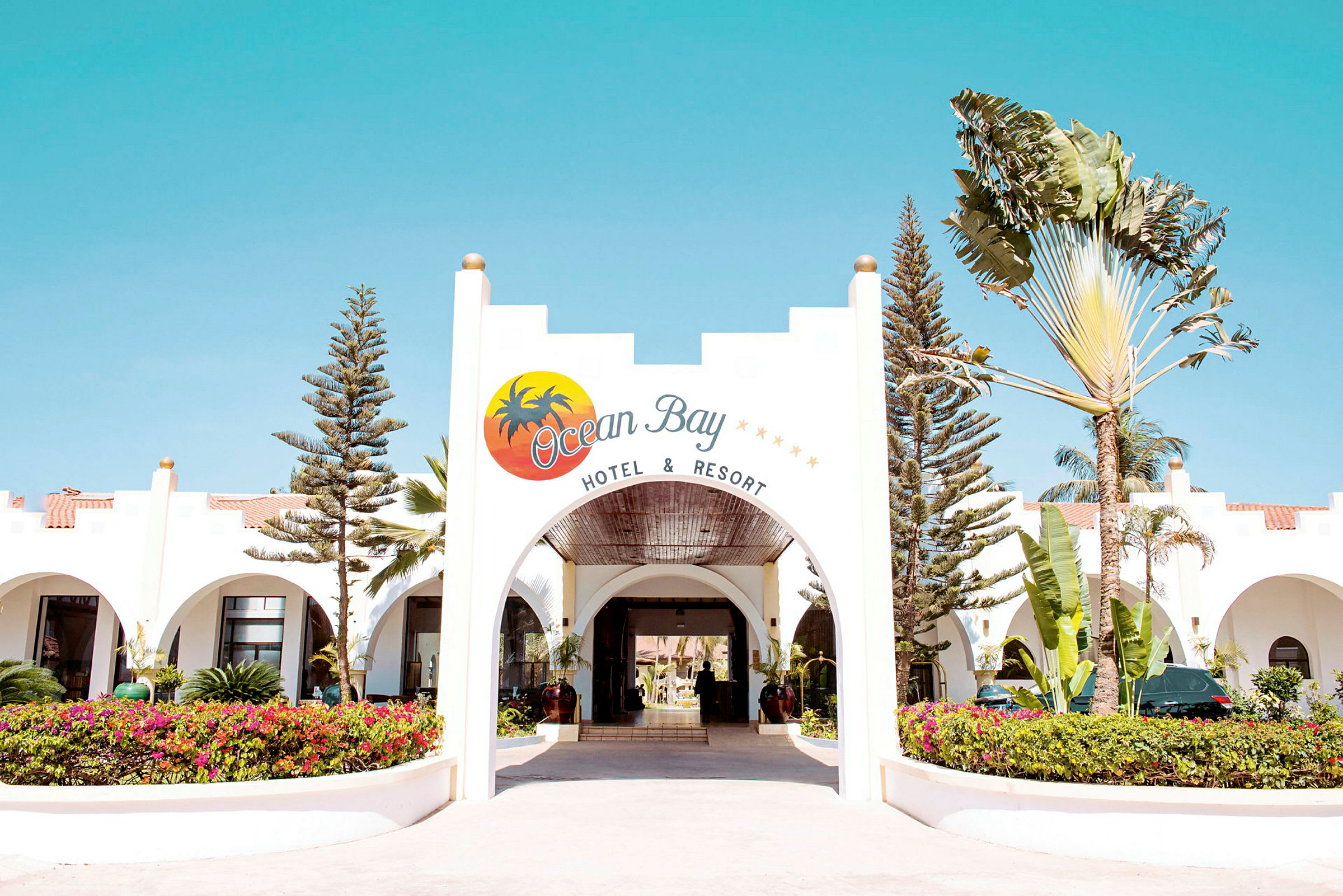 Indgangen til Ocean Bay Hotel & Resort, med en buet åbning og omkringliggende beplantning. (AI-genereret tekst)