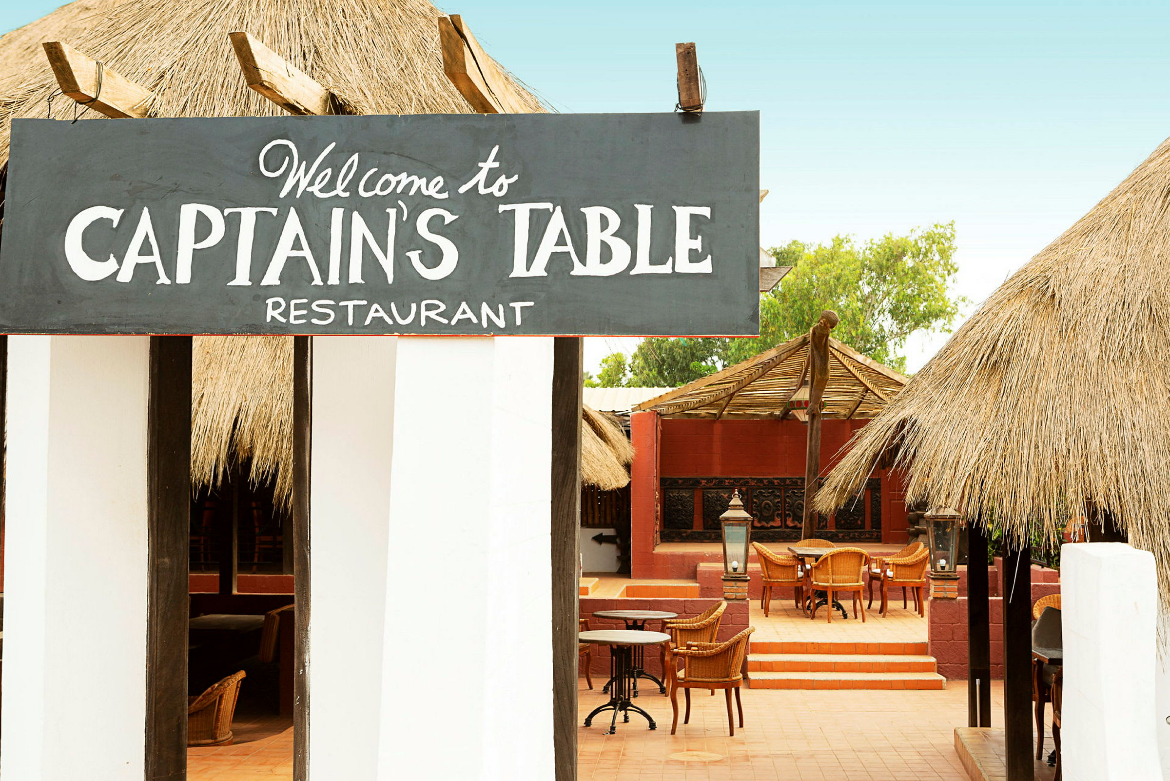 Et restaurantområde med skiltet "Welcome to Captain's Table Restaurant", stole og borde synlige udendørs under tag. (AI-genereret tekst)