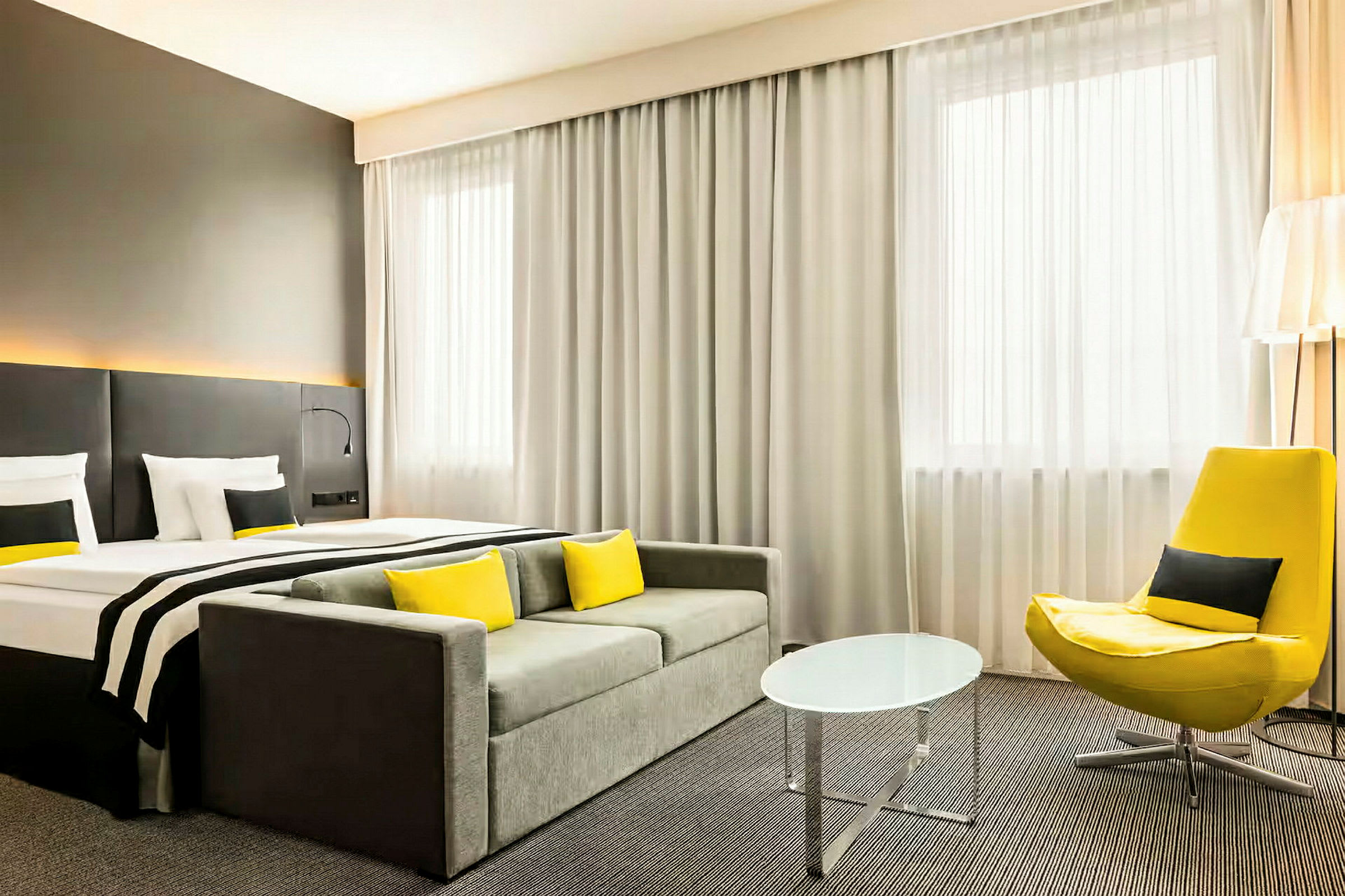 Et hotelværelse med en seng, sofa, gul lænestol og et lille rundt bord. (AI-genereret tekst)