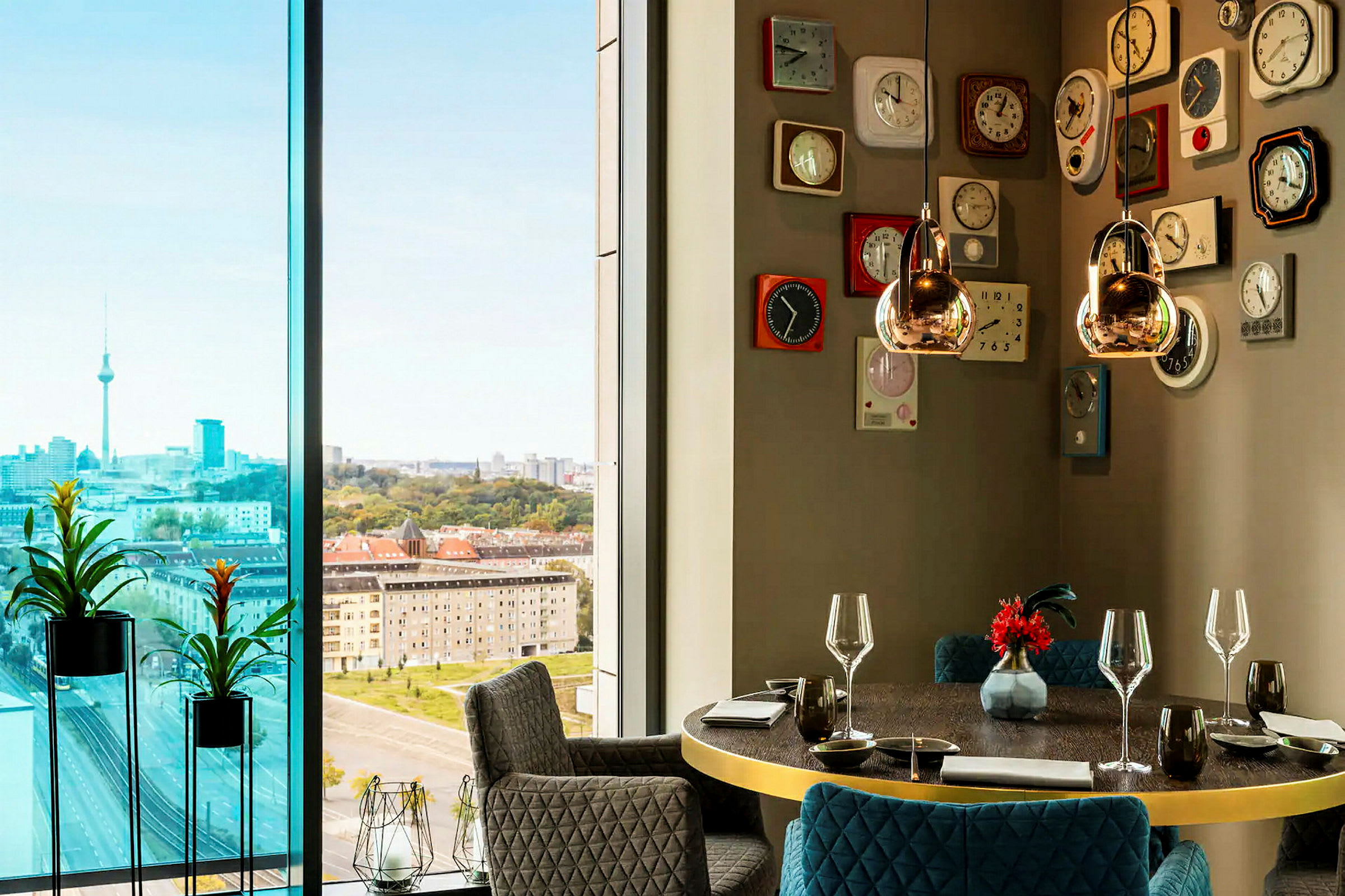 Et moderne rum med store vinduer, der viser en urban udsigt med et tårn omgivet af høje bygninger og grønne områder. Interiøret inkluderer et rundt spisebord med glas og porcelæn samt en væg dekoreret med retro-inspirerede ure. (AI-genereret tekst)