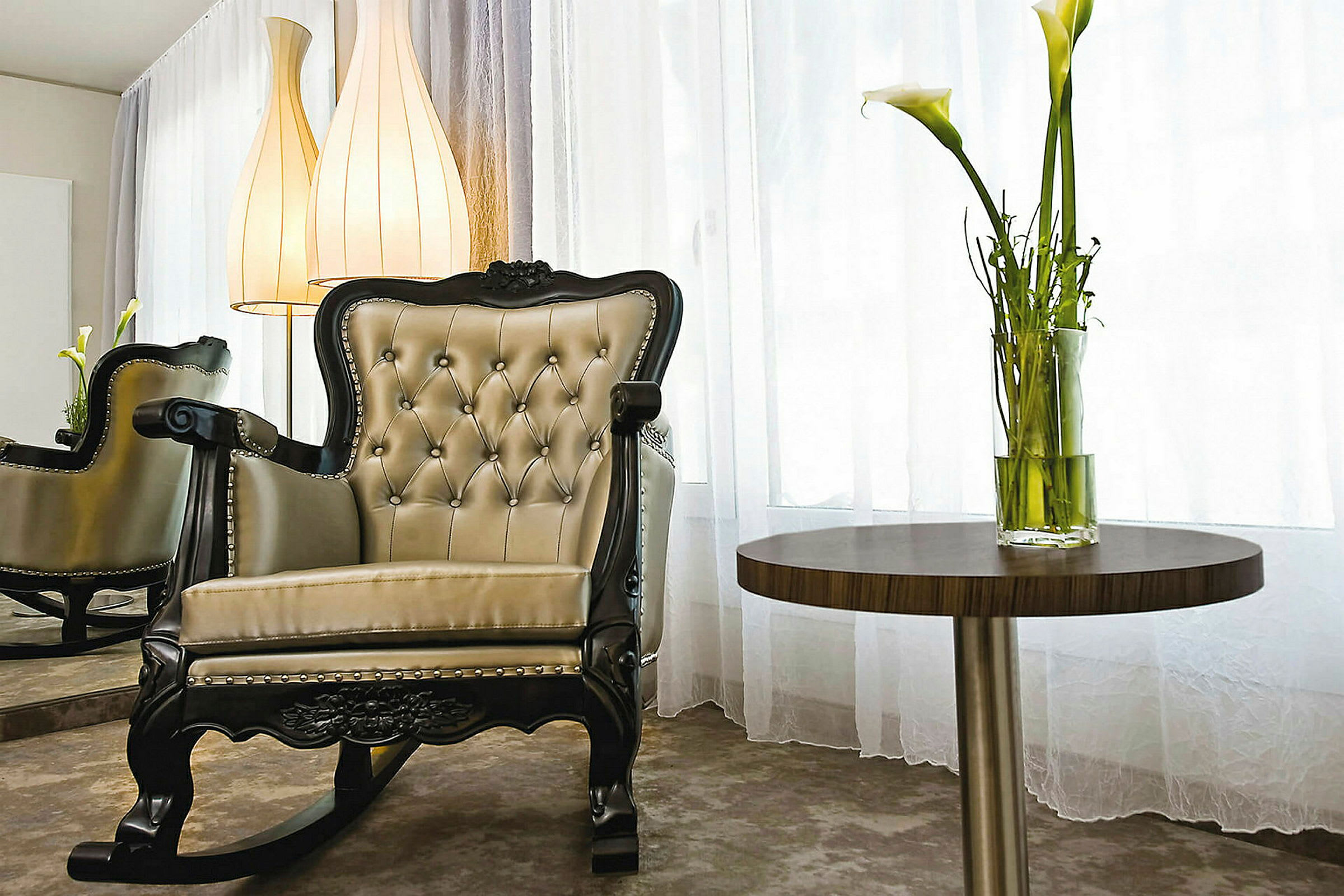 En elegant gyngestol med polstring og et rundt sidebord med en vase med hvide liljer, i et rum med lyse gardiner. (AI-genereret tekst)