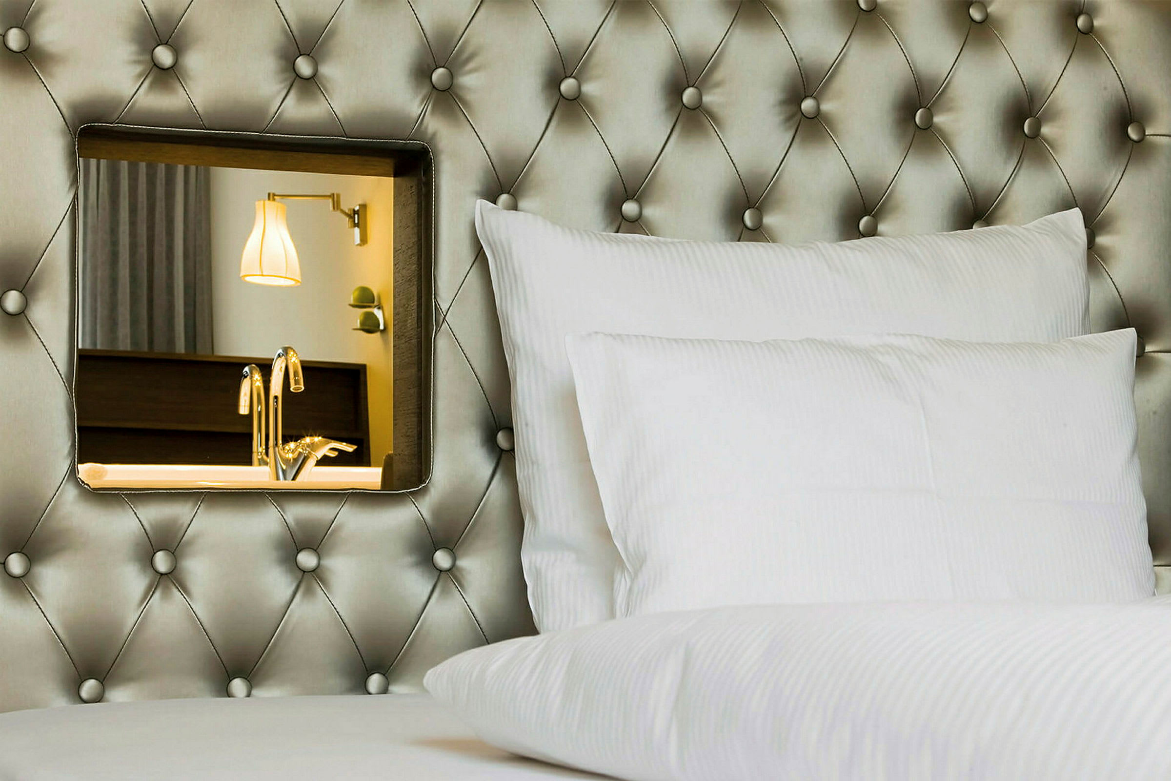 Hotelseng med hvid pude og dekorativt spejl på den polstrede sengegavl. (AI-genereret tekst)
