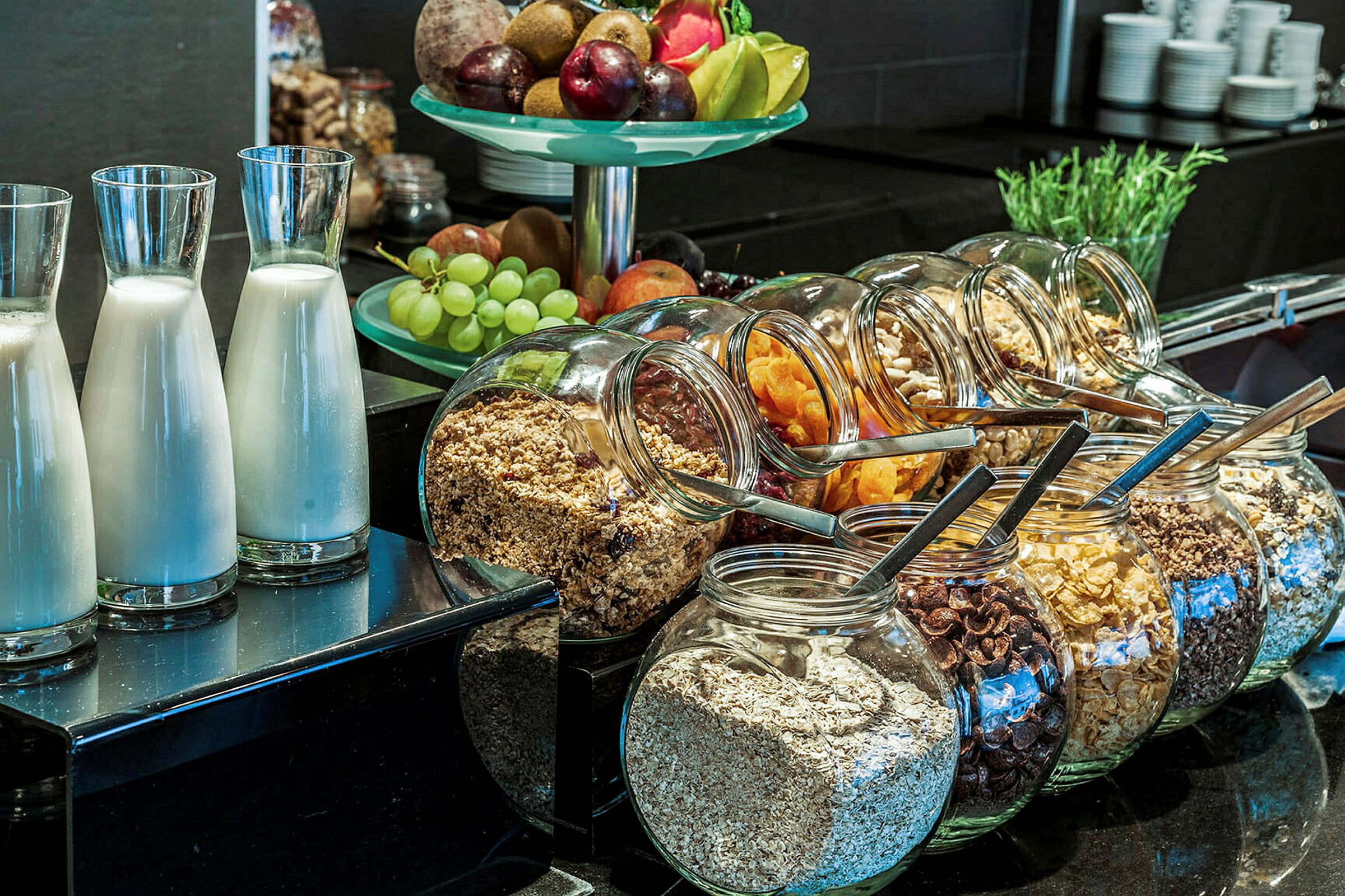En morgenbuffet med glasflasker med mælk, glasbeholdere med forskellige slags cornflakes og mysli samt en skål med frisk frugt. (AI-genereret tekst)