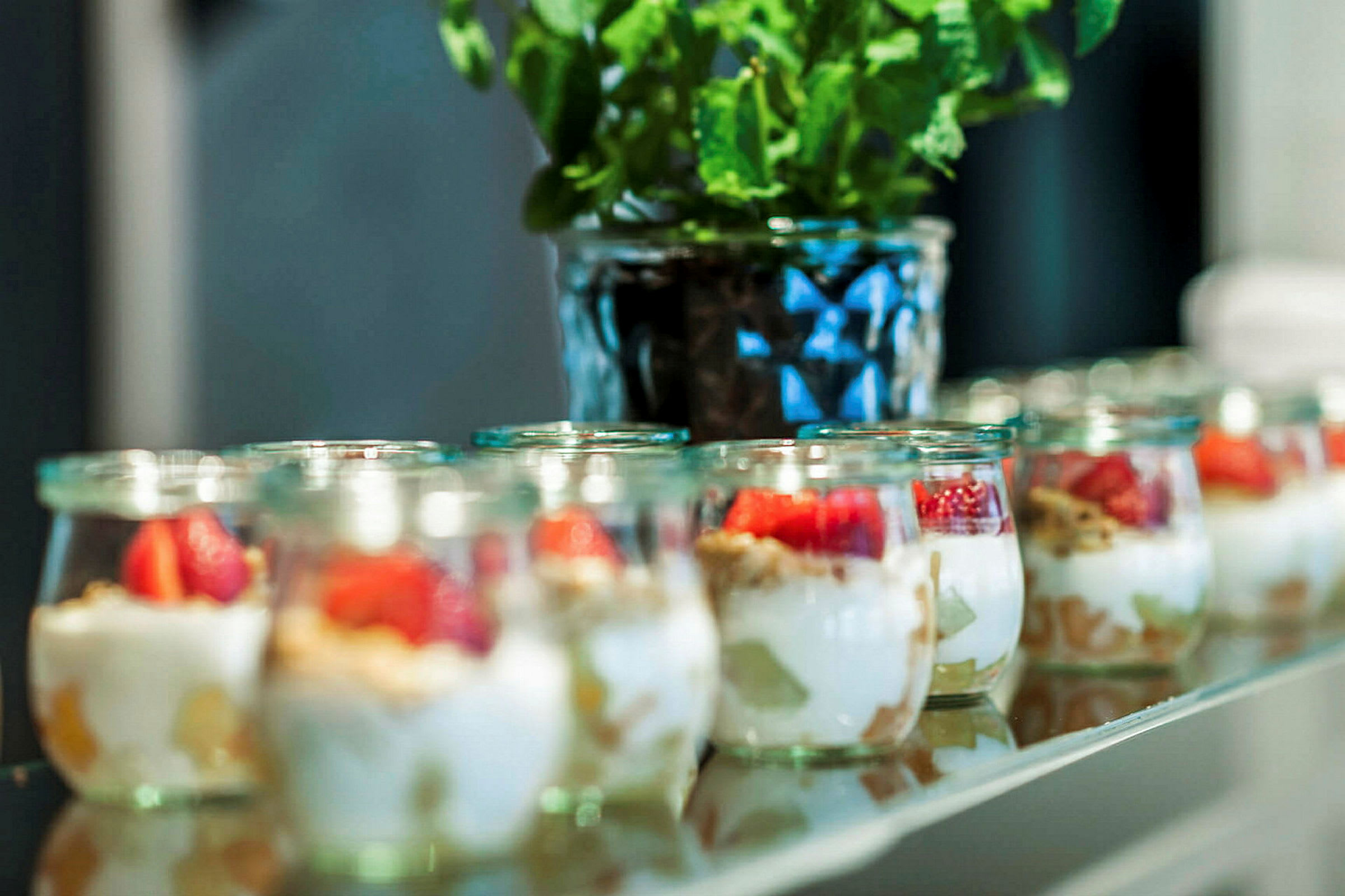 Små glasbeholdere med yoghurt, frugt og granola står på række. I baggrunden ses en potteplante. (AI-genereret tekst)